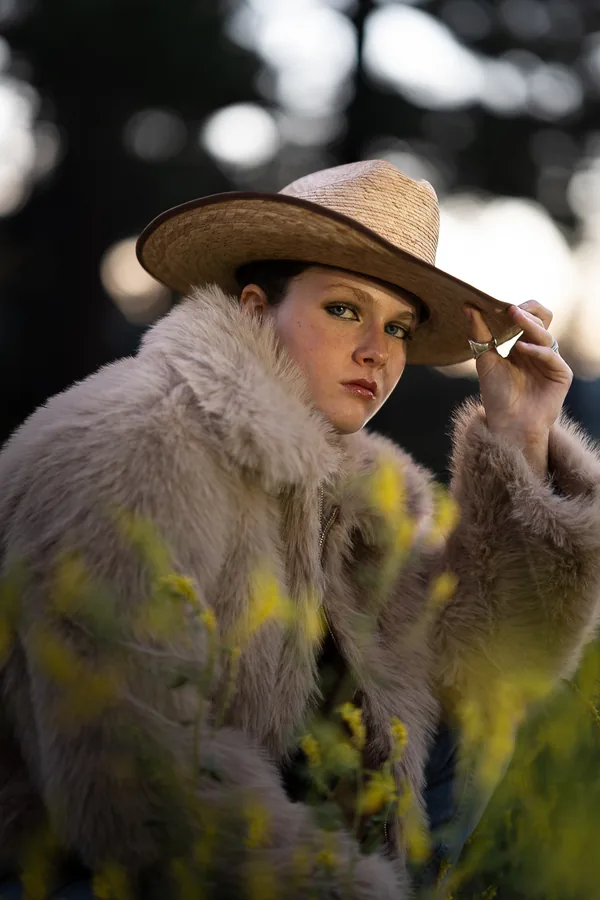 Woman in a straw cowboy hat and cream fur coat tipping the brim, framed through yellow wildflowers in warm backlight