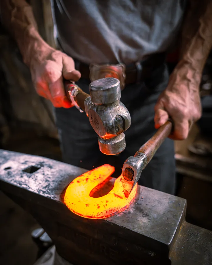 Blacksmith hammering a glowing orange horseshoe on an anvil in a dark forge
