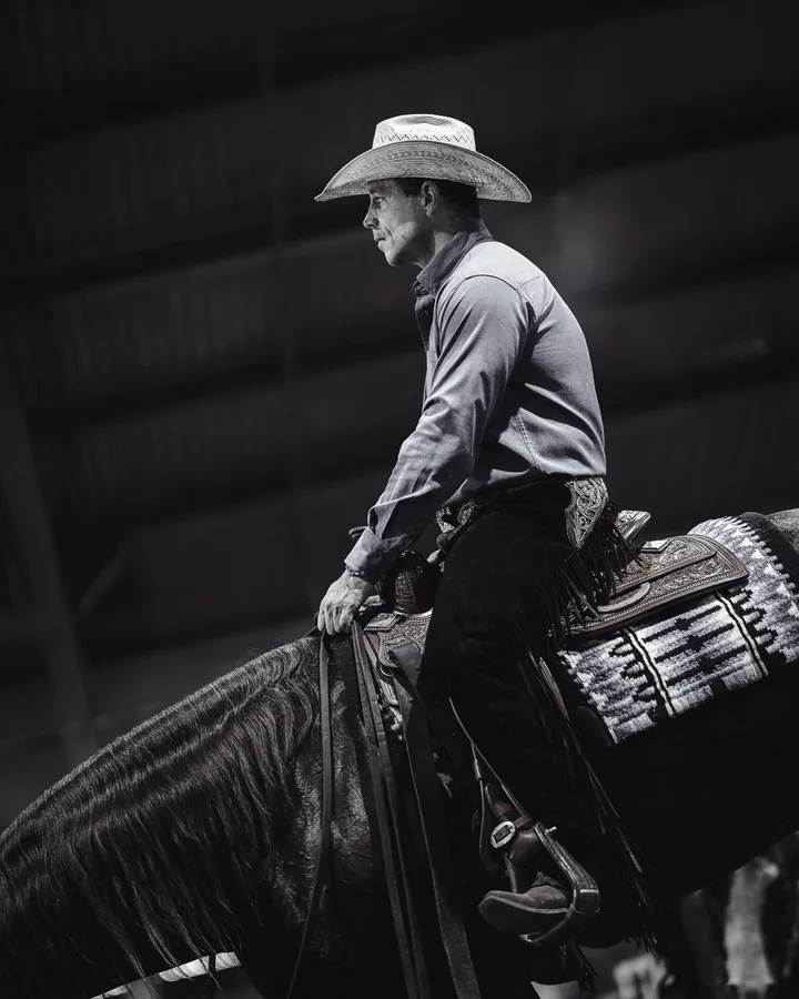 Black and white photo of a cowboy in chaps riding a dark horse across a dimly lit arena
