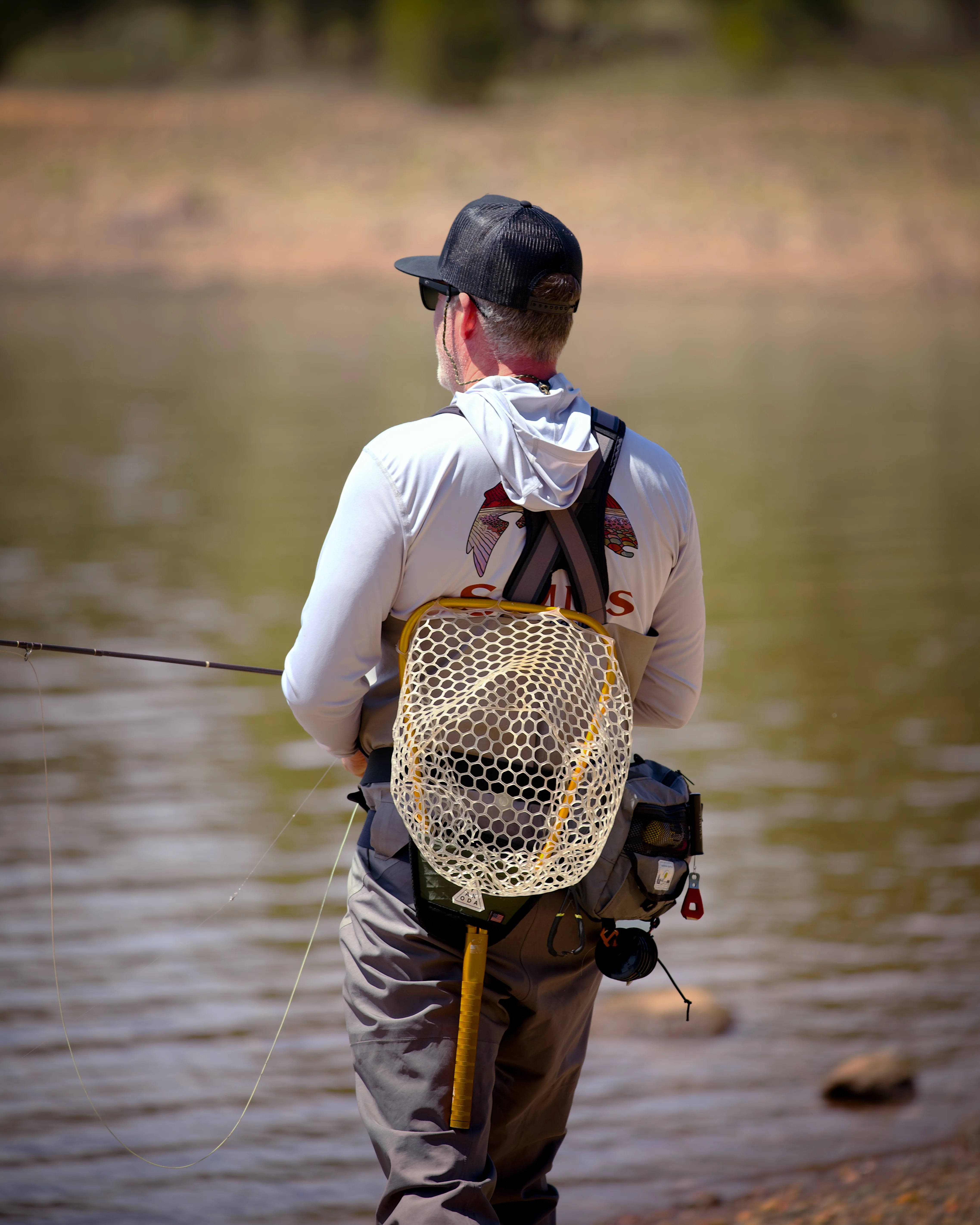Fly fisherman seen from behind on a riverbank in waders, holding a fly rod with a landing net on his back