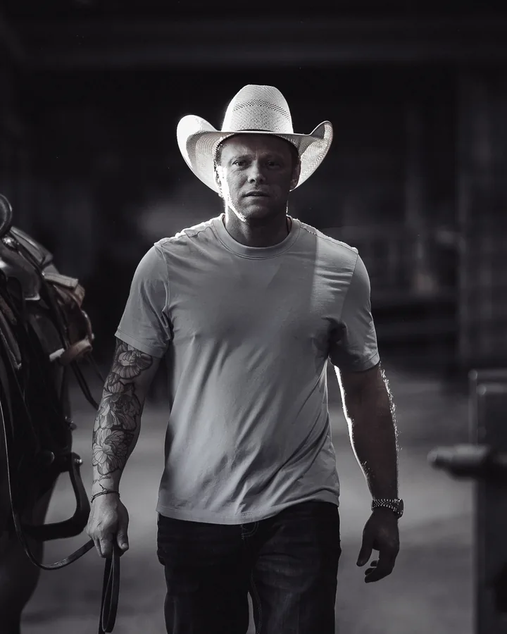 Black and white portrait of a cowboy in a straw hat and t-shirt leading a saddled horse through a barn aisle