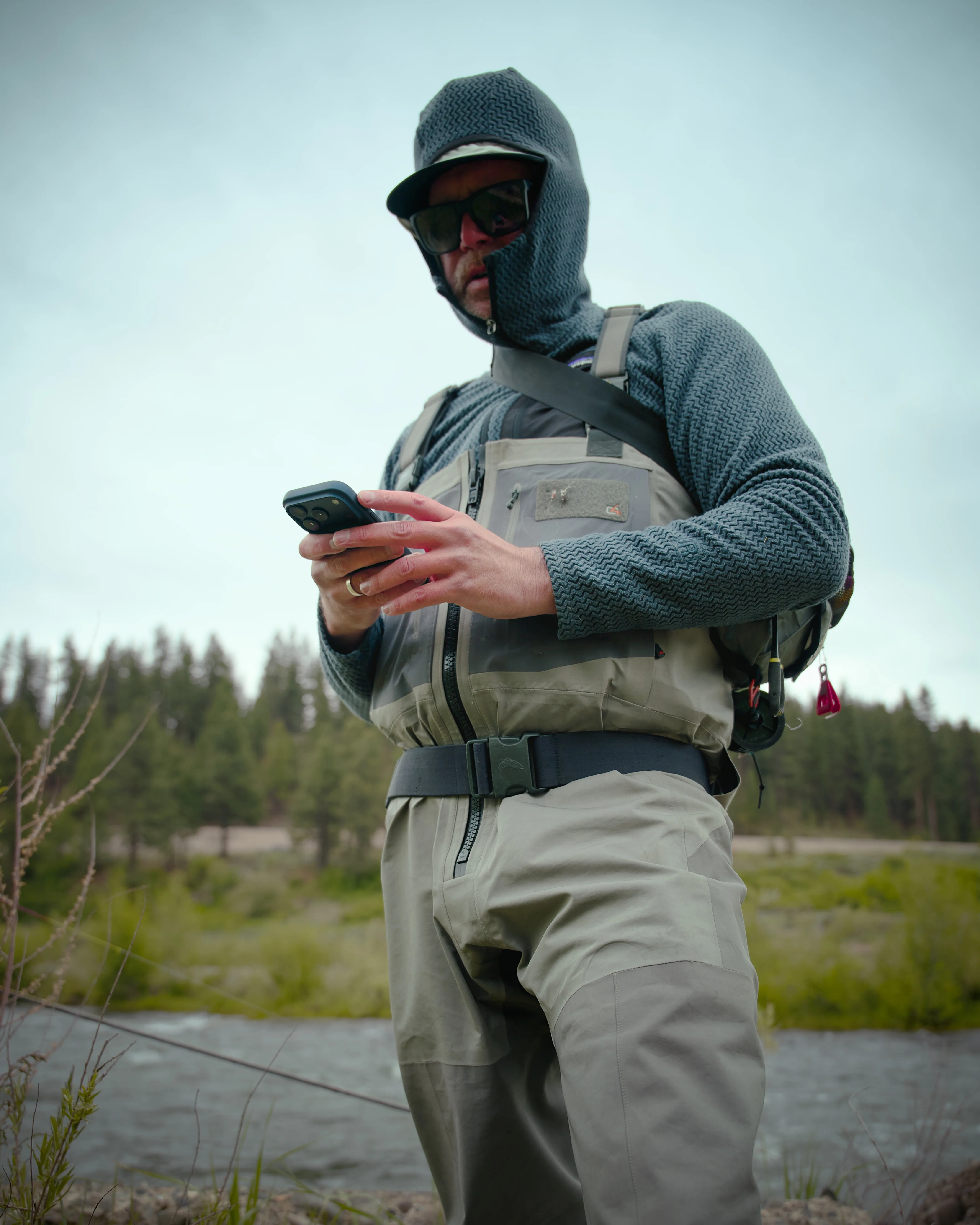 Fly fisherman in waders and a balaclava checking his phone beside a river with a pine forest in the background