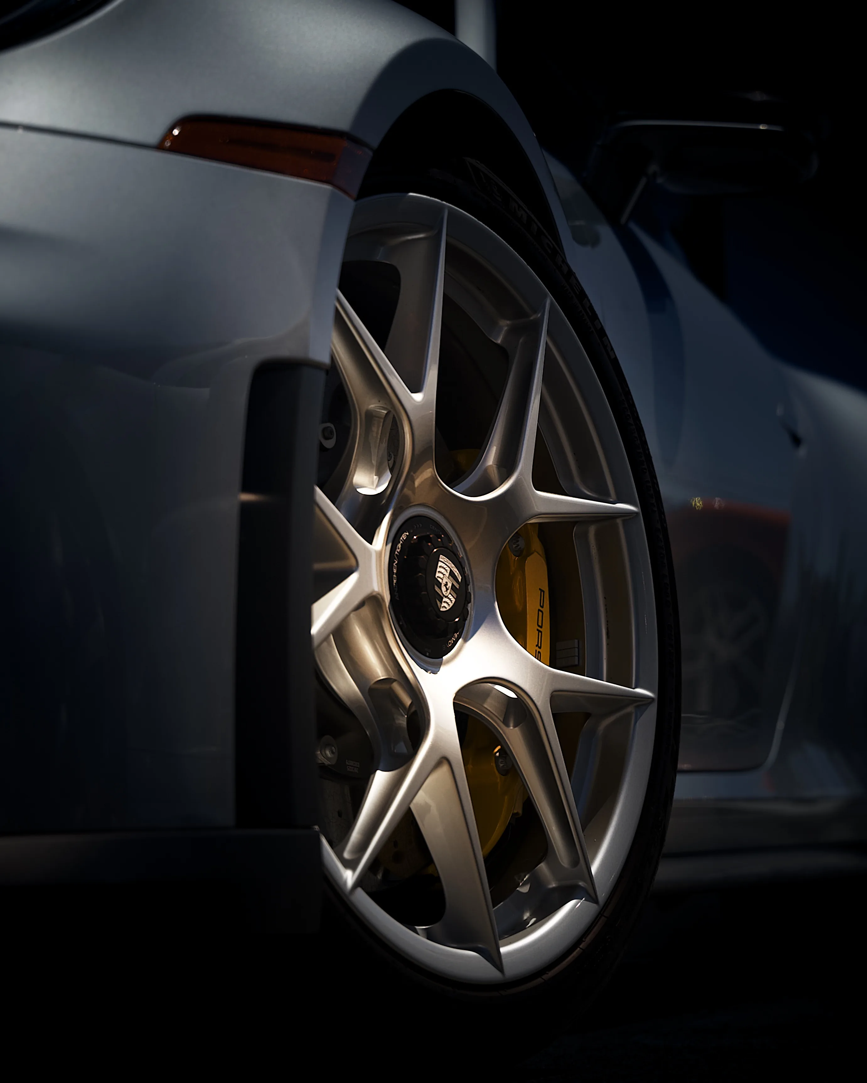 Close-up of a silver Porsche wheel with yellow brake calipers and a Porsche center cap, lit dramatically against a dark background