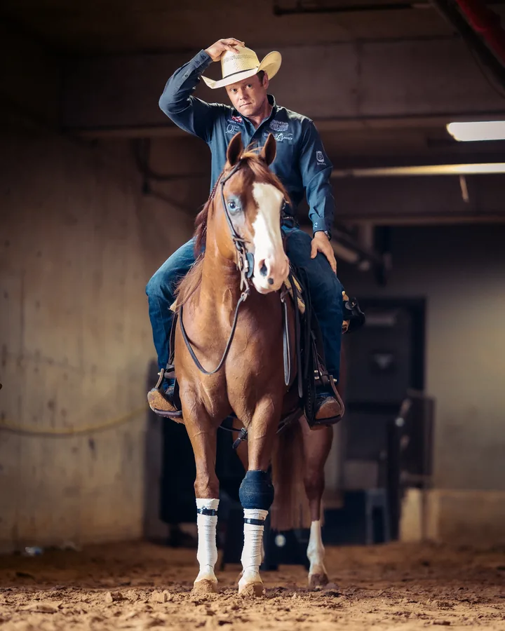Cowboy in a navy shirt tipping his straw hat while riding a chestnut horse with a white blaze through a warmly lit barn