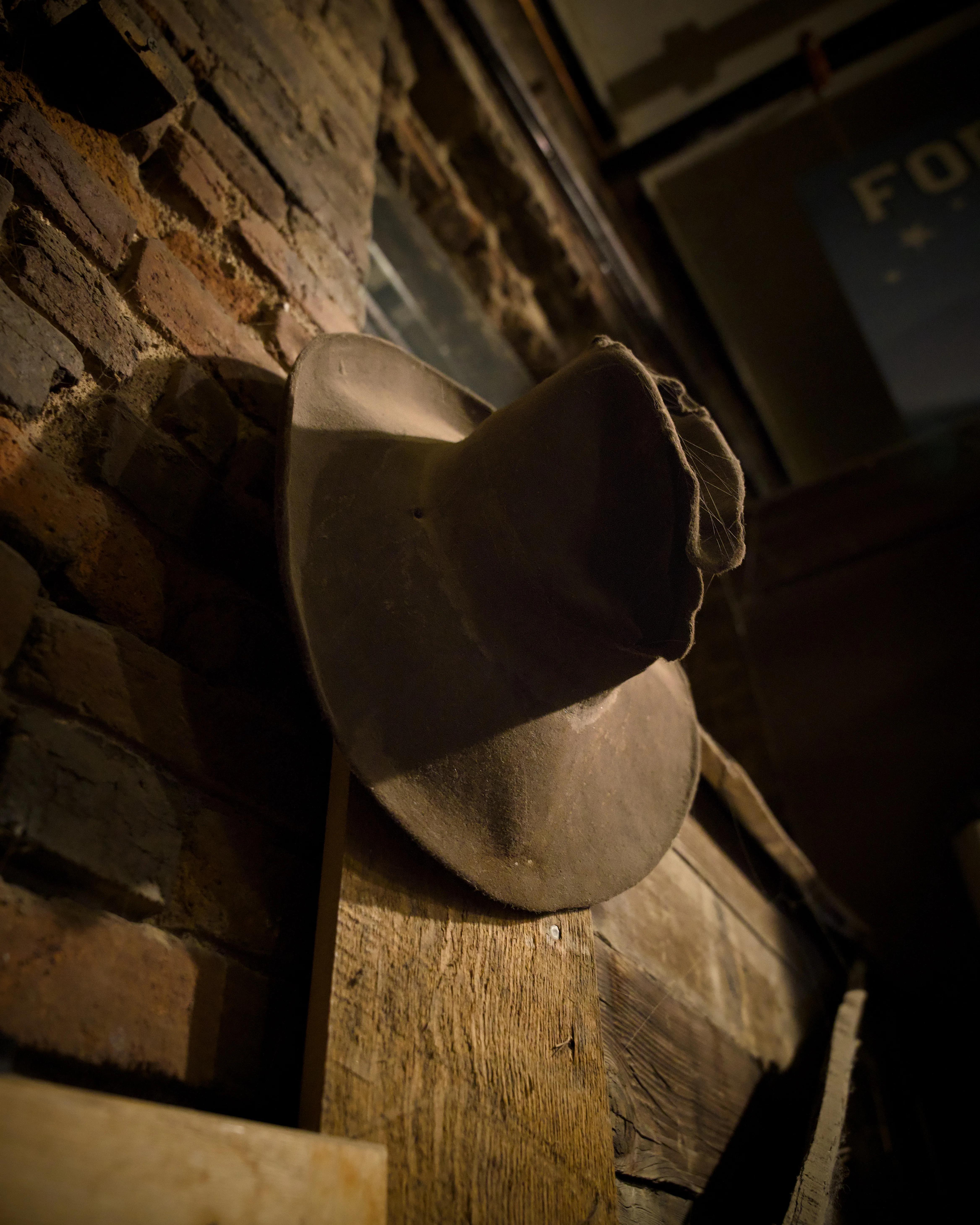 Worn grey felt cowboy hat resting on a rough wooden beam against a rustic brick wall in dim moody light