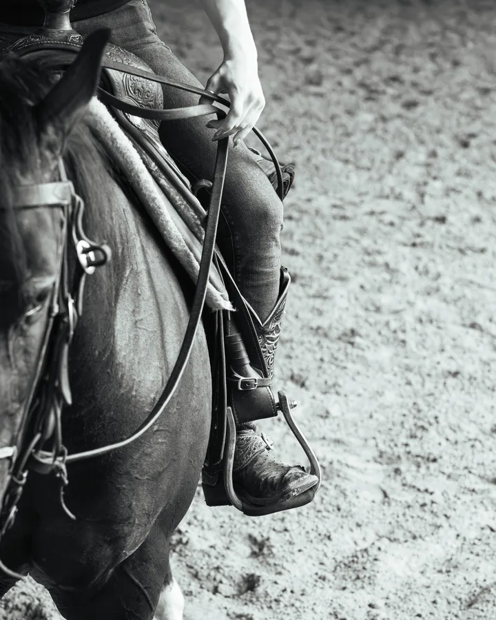 Black and white close-up of a cowboy boot in a stirrup with tooled leather chaps and reins draped over a horse's side