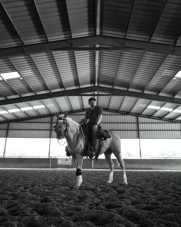 Black and white photo of a rider on a light grey horse standing in the center of a covered arena