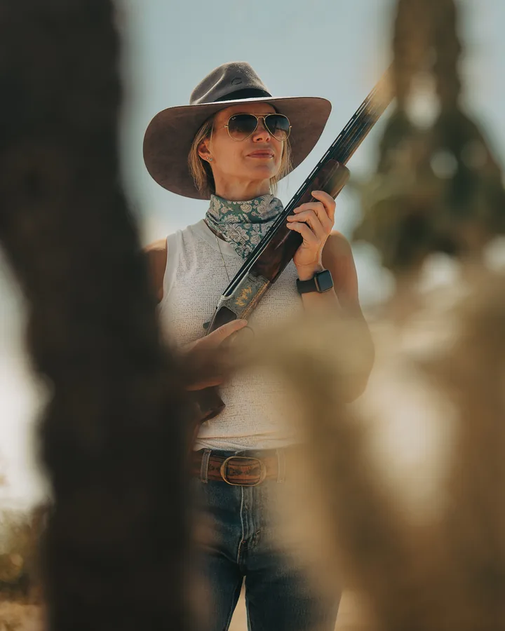 Woman in a felt hat and sunglasses holding a shotgun upright in warm desert light, framed by out-of-focus branches