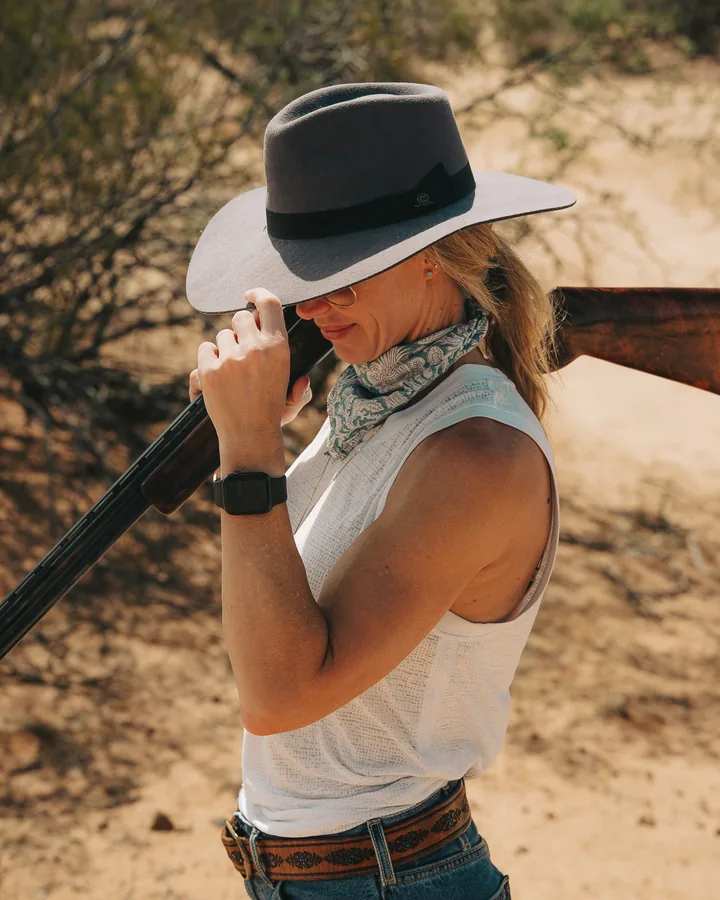 Woman in a grey wide-brim hat and paisley bandana resting a shotgun on her shoulder in the desert