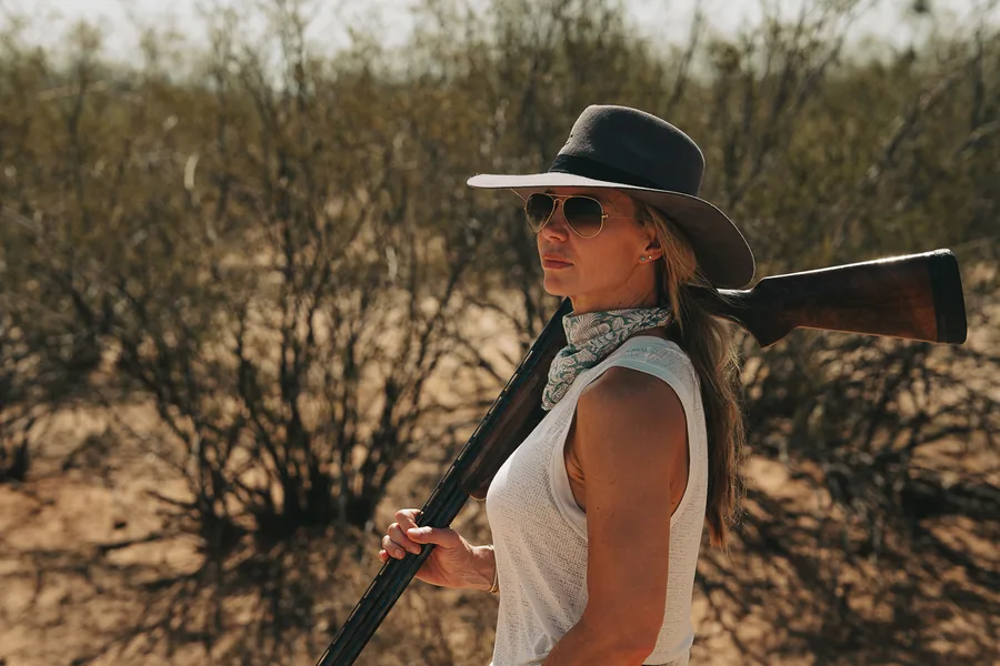 Woman in a black hat and sunglasses carrying a shotgun over her shoulder through sunlit desert brush