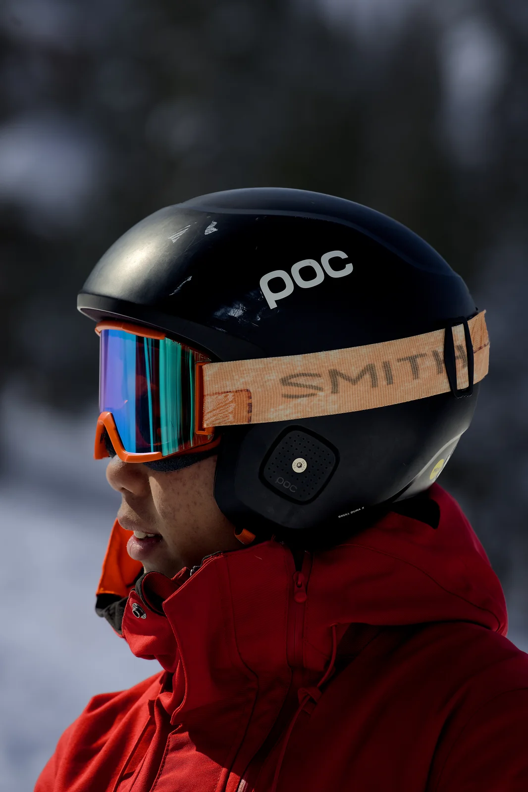 Skier in a red jacket wearing a black POC helmet and Smith goggles with a snowy forest in the background