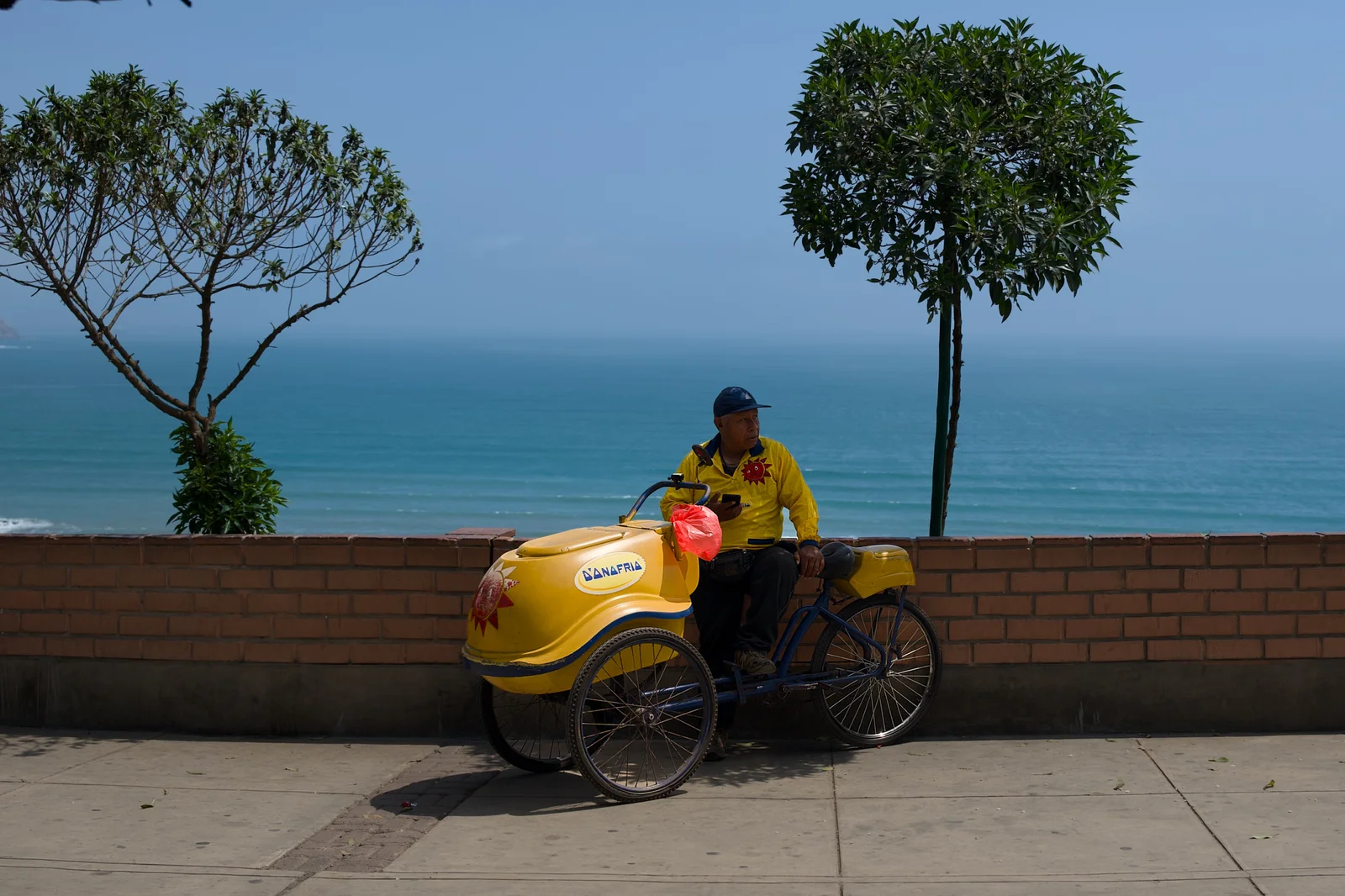Yellow food vendor on a bicycle cart parked along a low wall overlooking a bright turquoise ocean on a sunny day