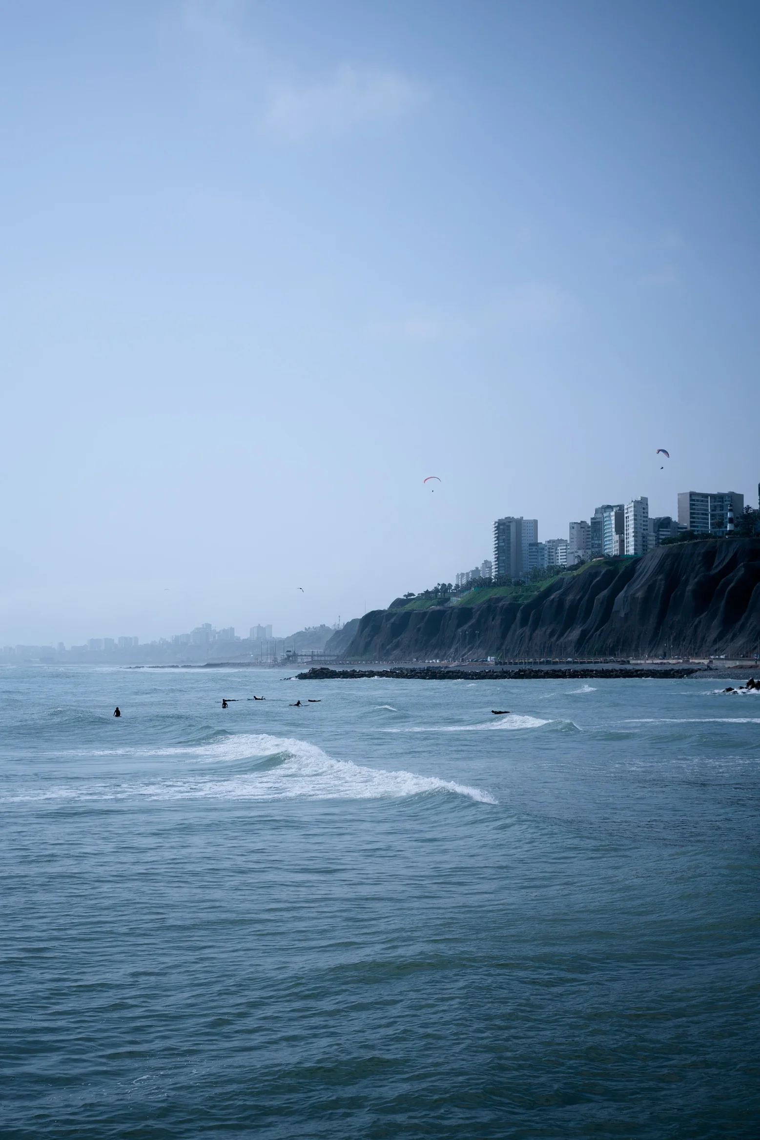Hazy coastal view of surfers in the water below green cliffs lined with buildings, paragliders drifting in a pale blue sky