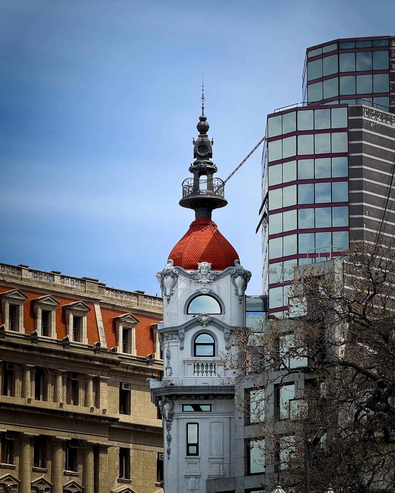 Ornate historic building tower with a red domed roof and decorative iron spire, framed between modern glass skyscrapers