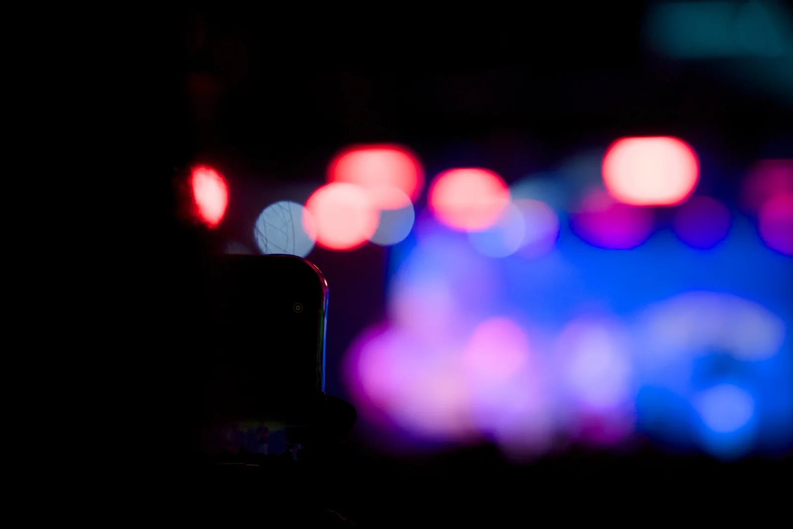 Silhouetted phone screen against blurred bokeh of red, pink, and blue concert stage lights