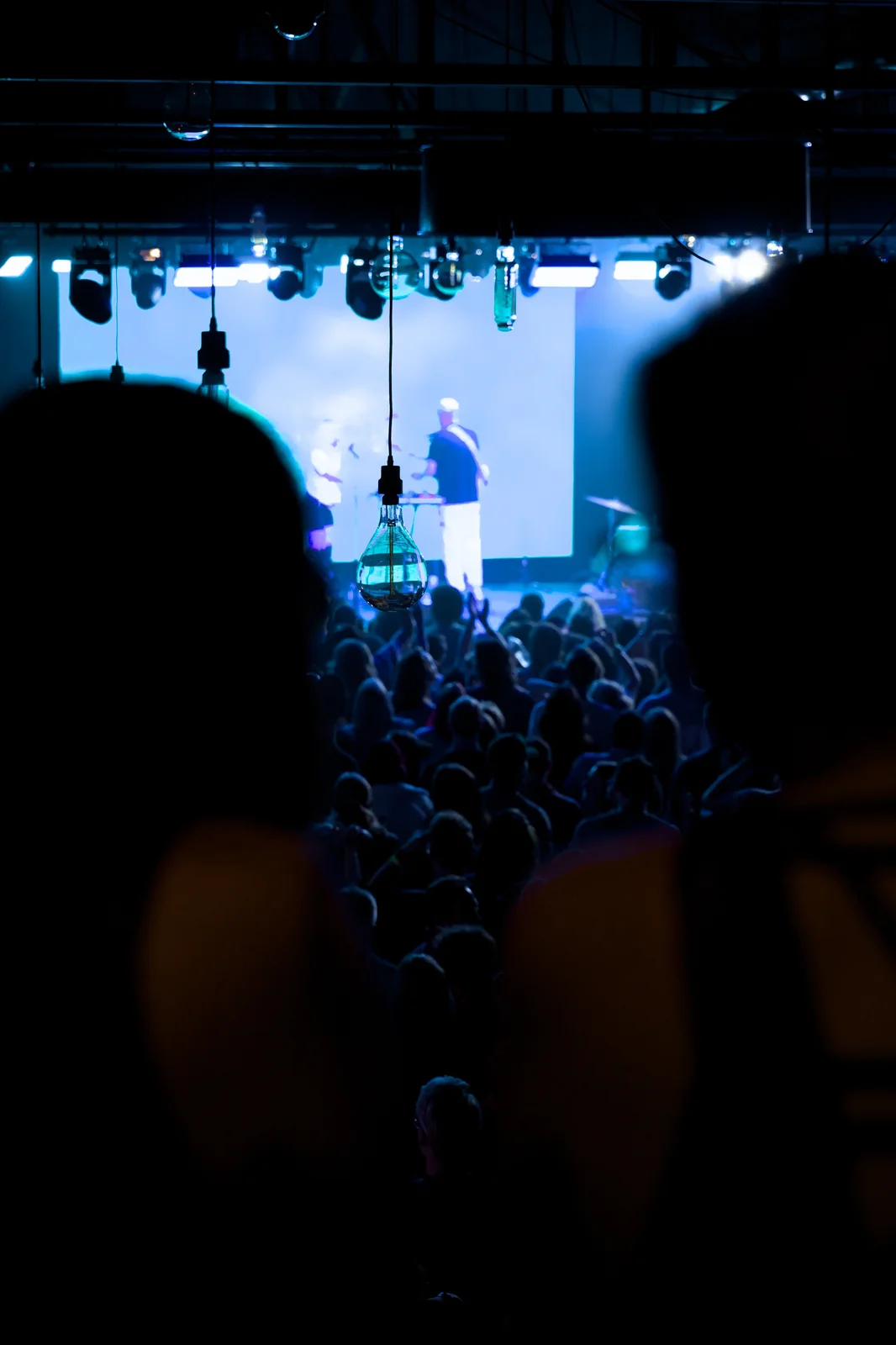 Wide shot of a packed concert crowd from behind with a performer on a blue-lit stage ahead