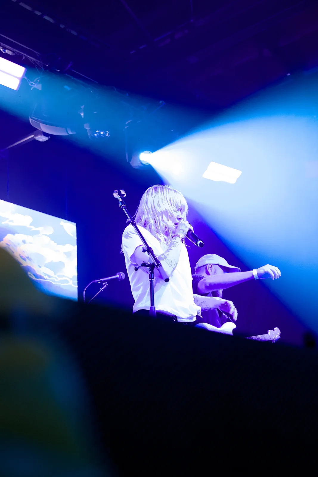 Female vocalist with pink-white hair singing into a microphone under blue-purple stage lights