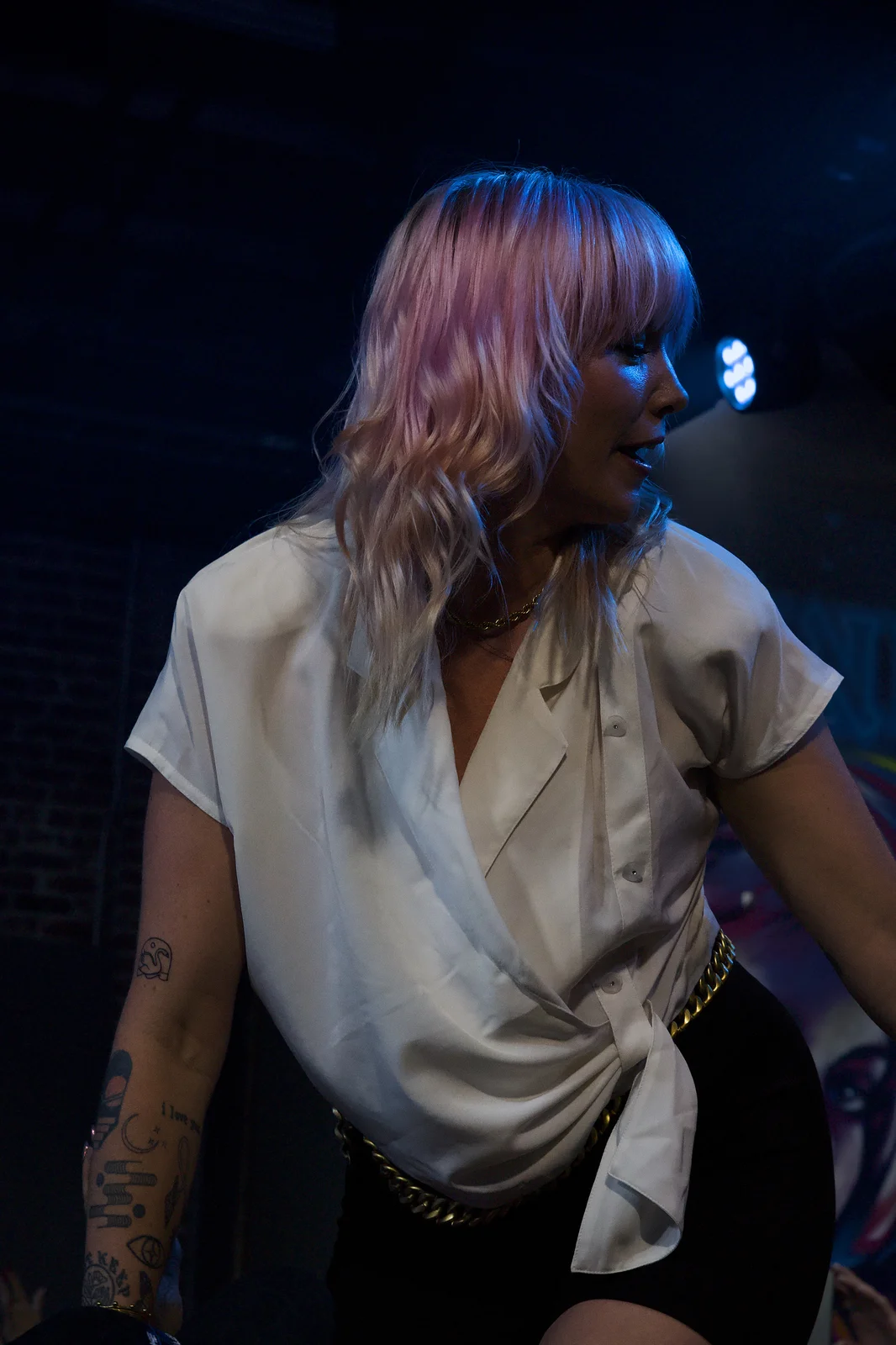 Female performer with pink hair leaning forward on stage in a white top under dark blue lighting