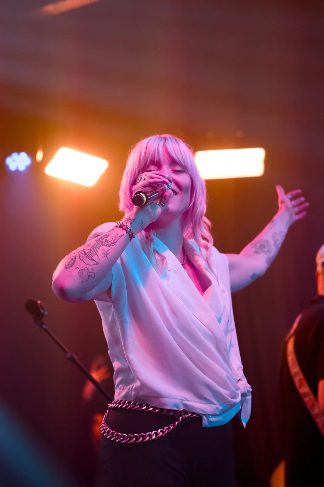 Pink-haired vocalist in a white blouse singing and gesturing on stage under warm orange lights