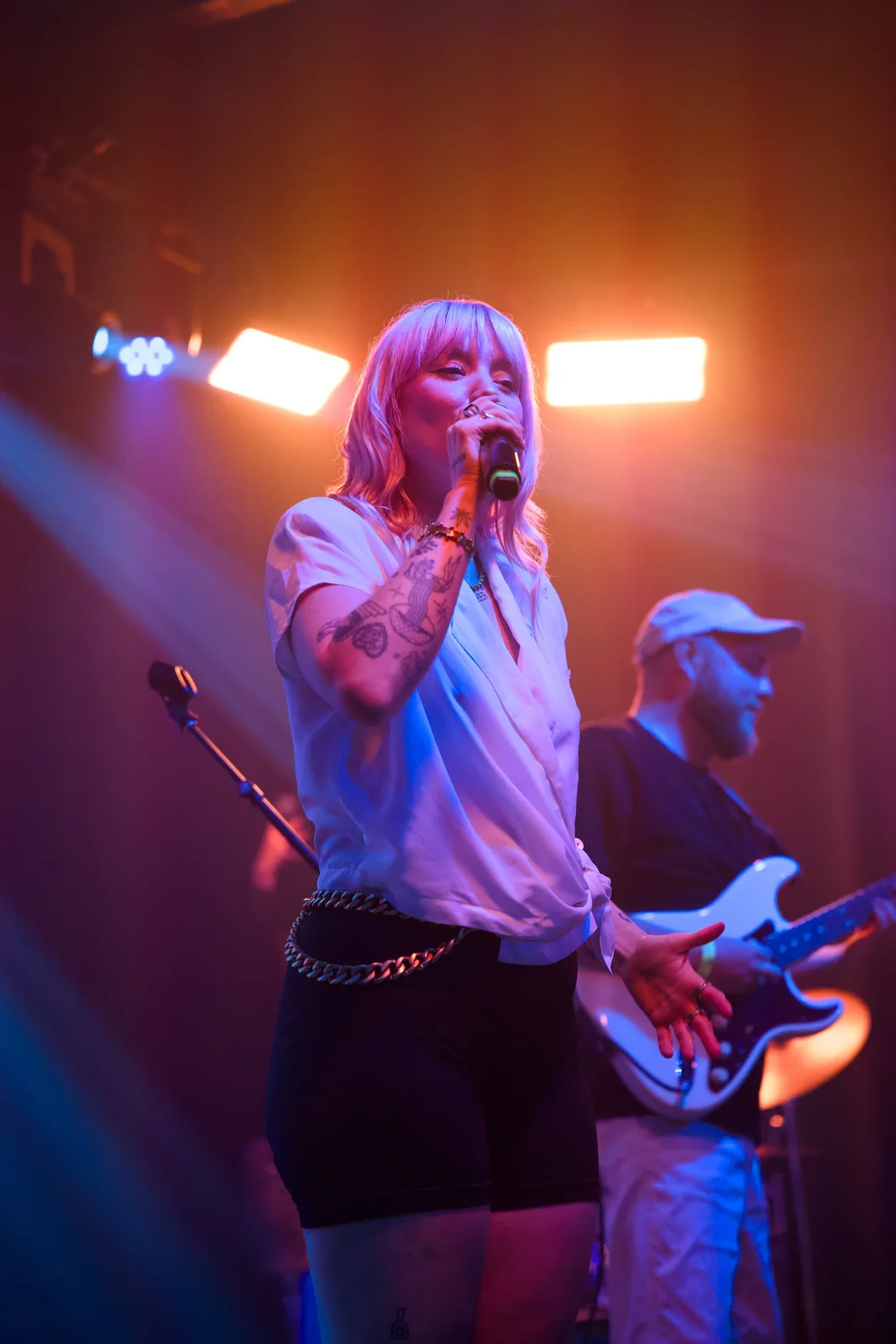 Female vocalist in a white top singing on stage with a guitarist in a white cap behind her