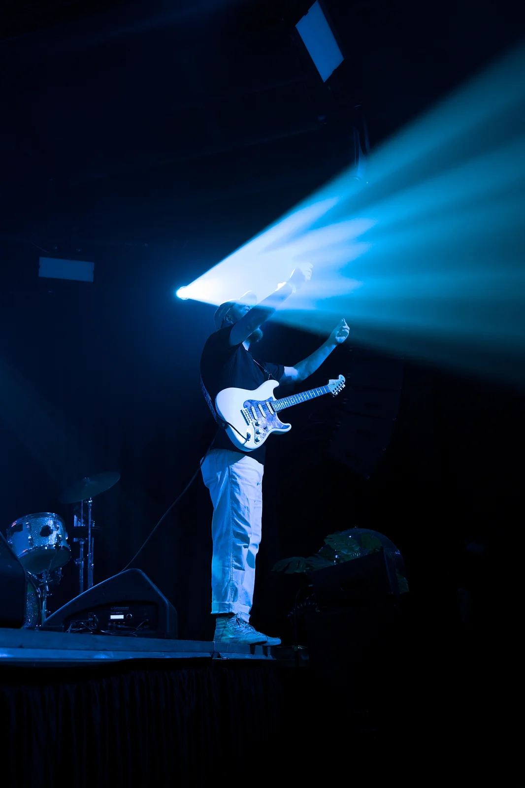 Male guitarist in white holding a Stratocaster raised overhead under a dramatic blue spotlight