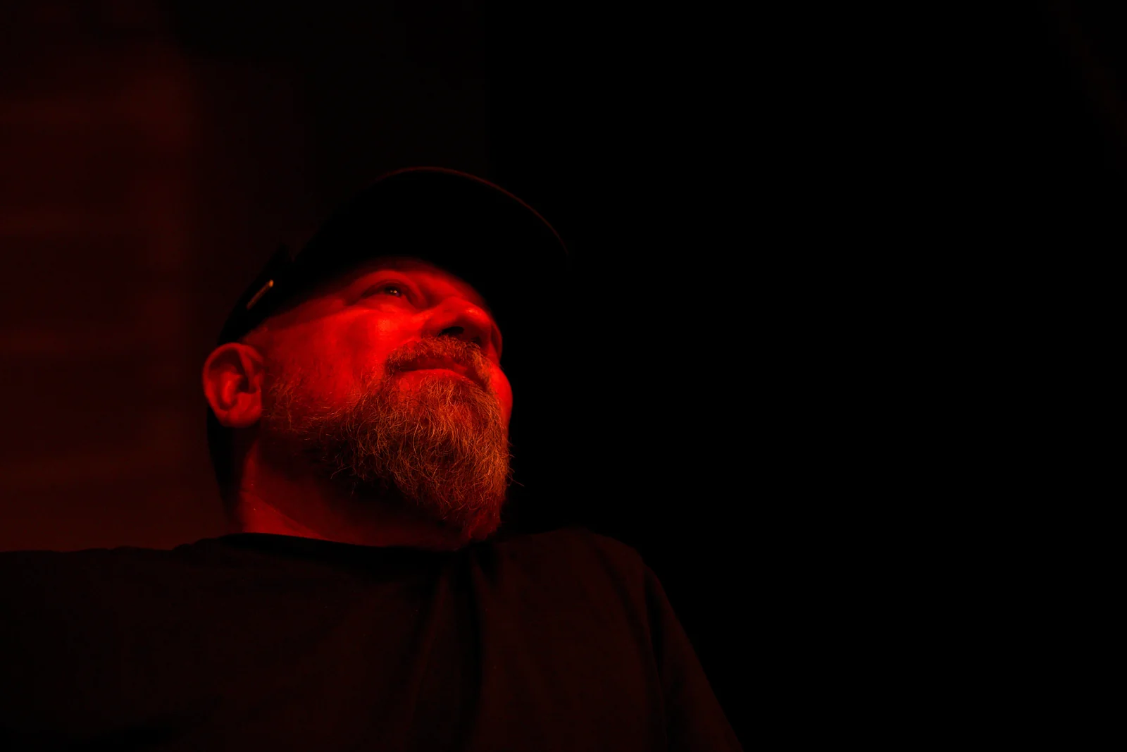 Bearded male musician illuminated by red stage light, gazing upward in a dark venue