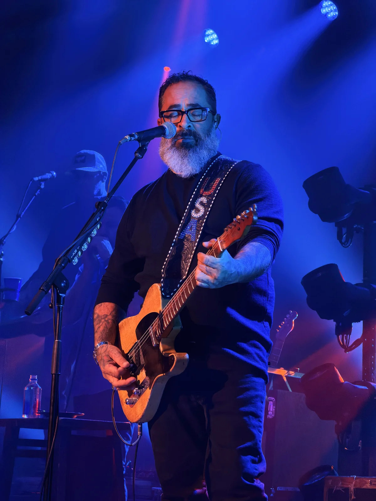 Bearded male guitarist with glasses singing and playing an acoustic guitar under blue stage lights