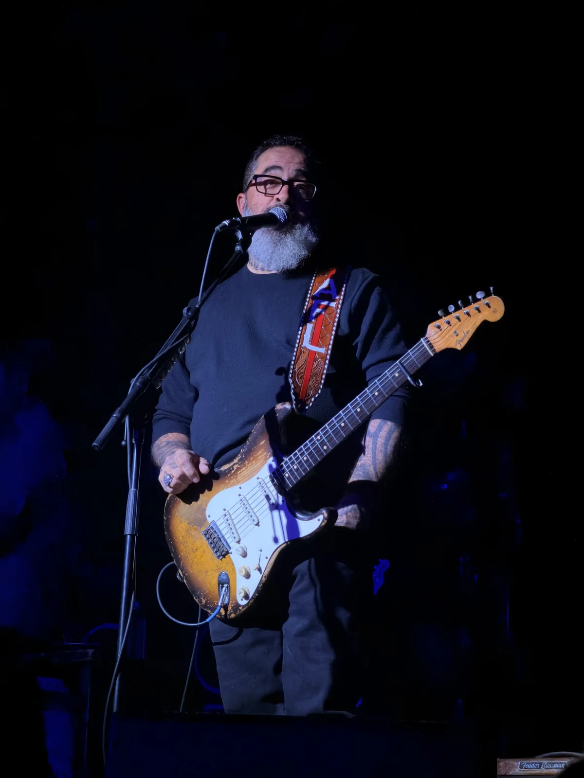 Bearded male guitarist with glasses singing at a mic while playing a battered sunburst Stratocaster