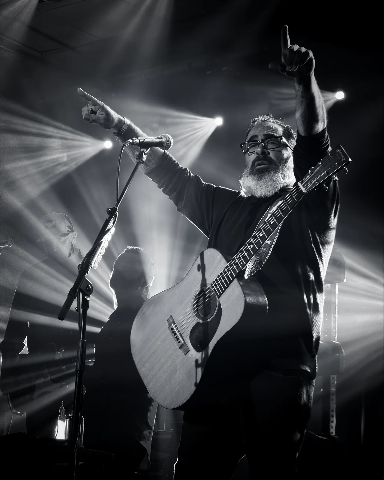 Bearded male guitarist with glasses raising both arms with an acoustic guitar under dramatic concert spotlights