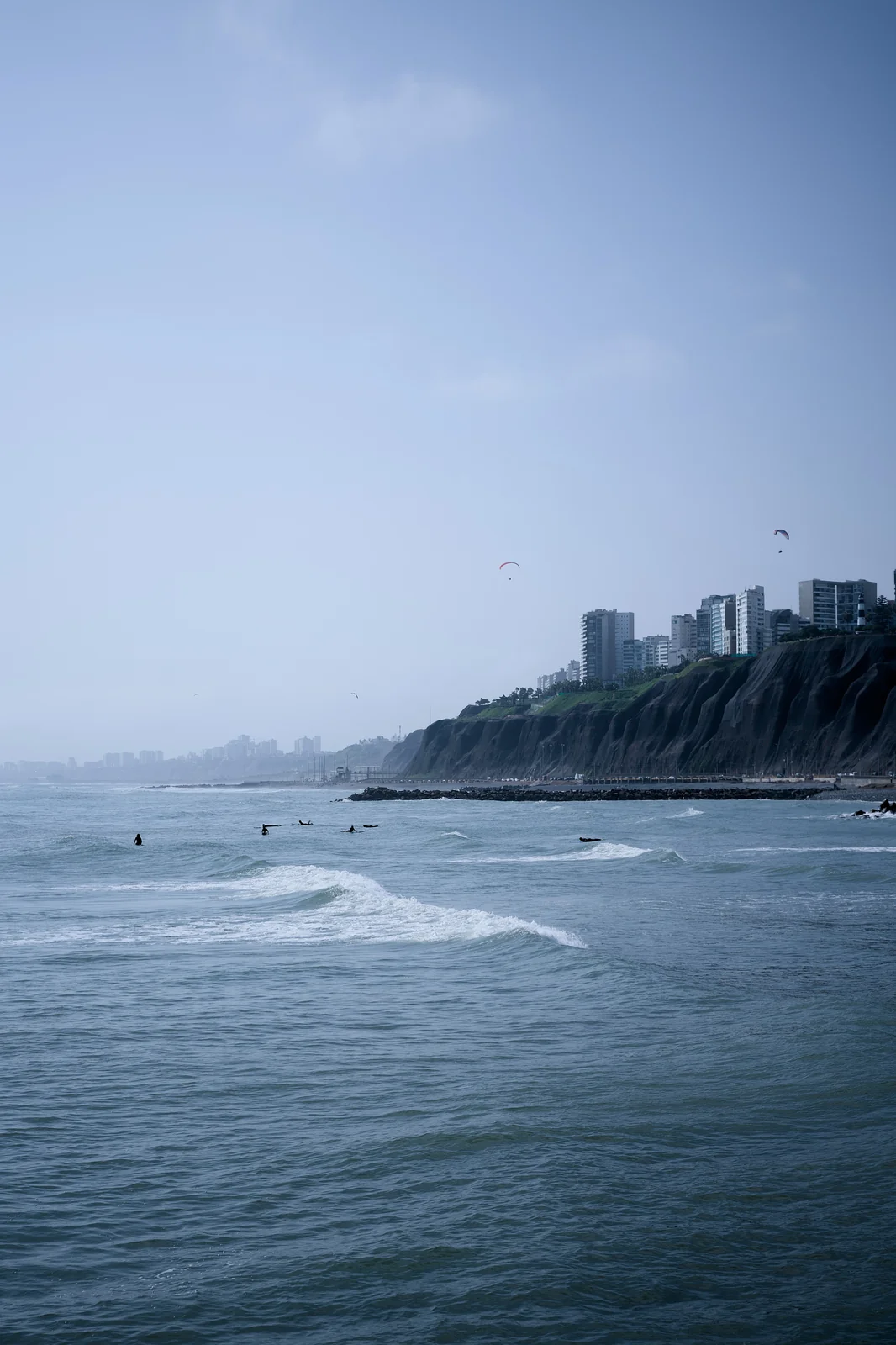 Coastline of Miraflores, Lima with surfers in the water, paragliders overhead, and city cliffs