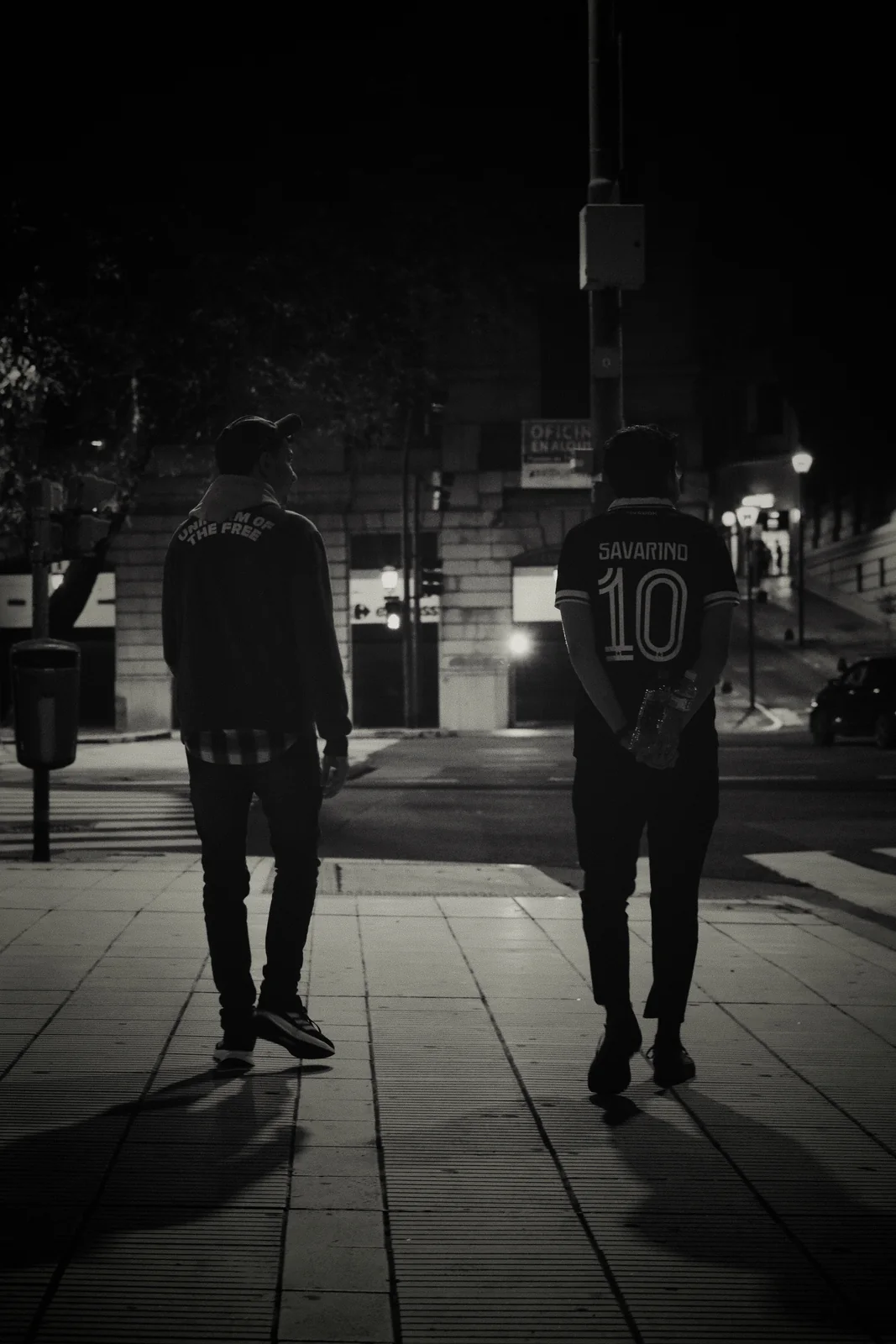 Two men walking down a city sidewalk at night in black and white, one wearing a Savarino jersey