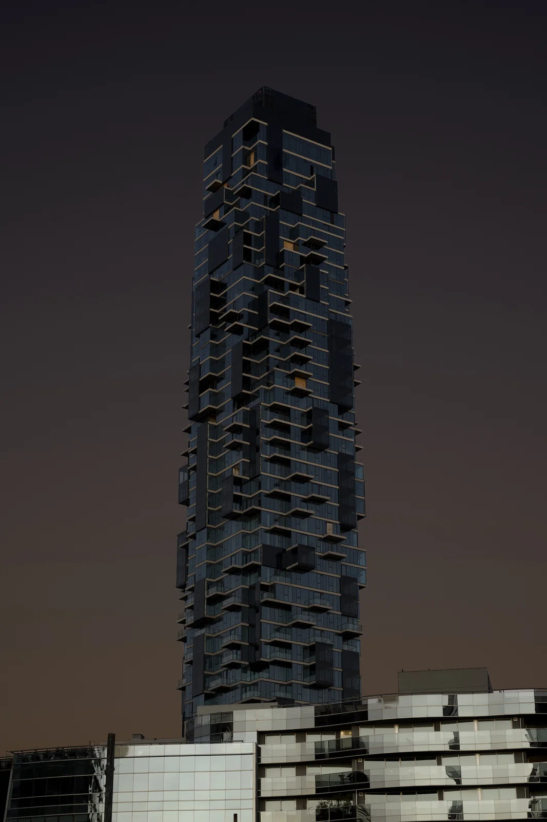 Tall glass skyscraper with staggered balconies rising against a dark moody dusk sky