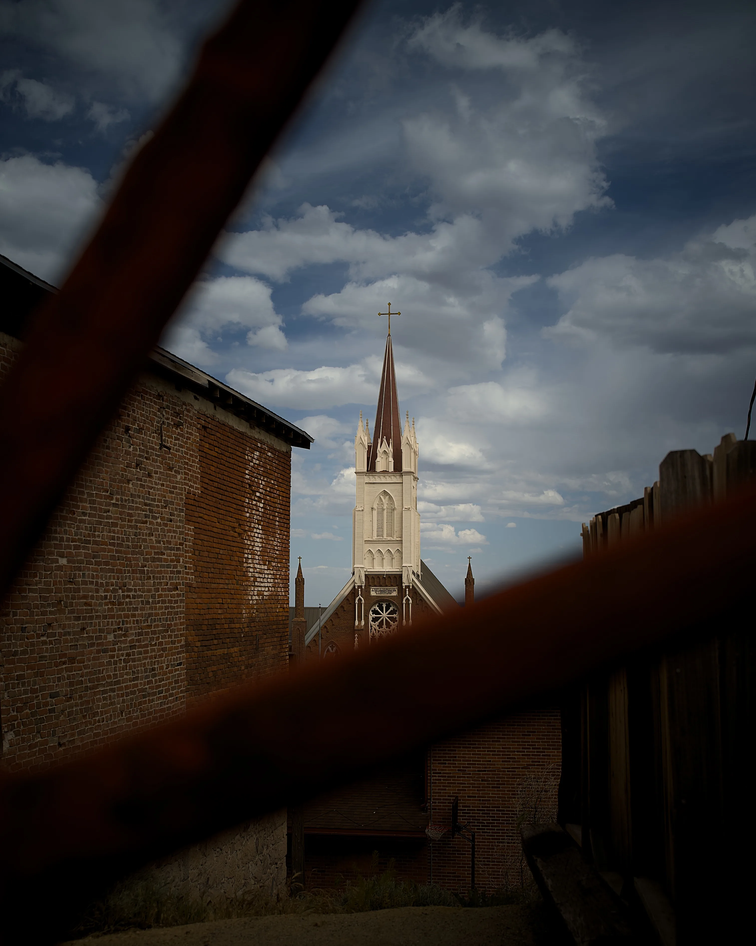 White Gothic church steeple with a gold cross rising above brick buildings, framed through rusty iron bars against a dramatic cloudy sky
