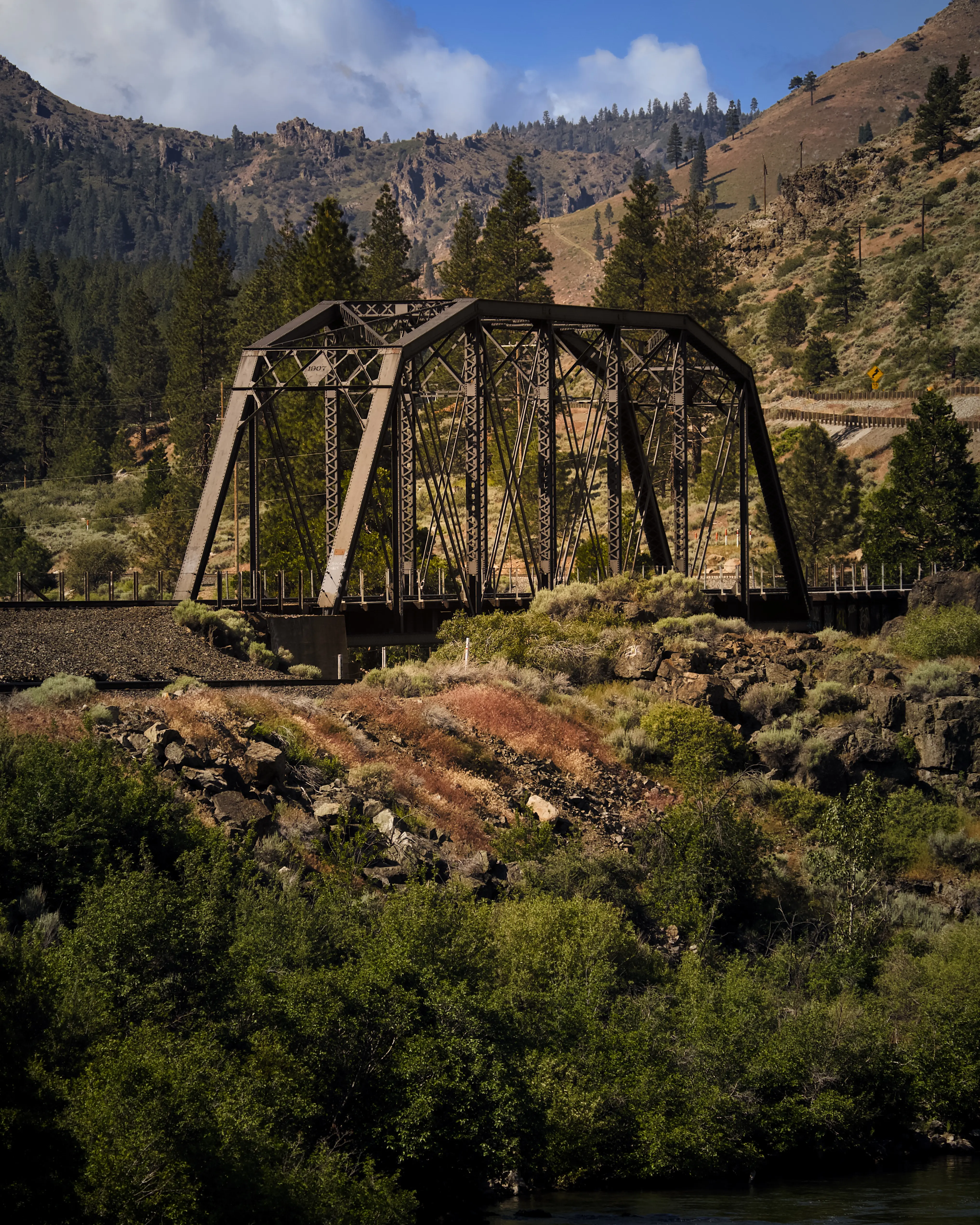 Rustic iron truss railroad bridge spanning a river canyon with pine-covered mountains and a partly cloudy sky