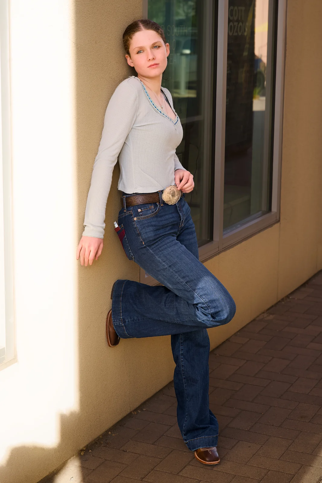 Woman in a grey top and flare jeans leaning against a building wall outdoors