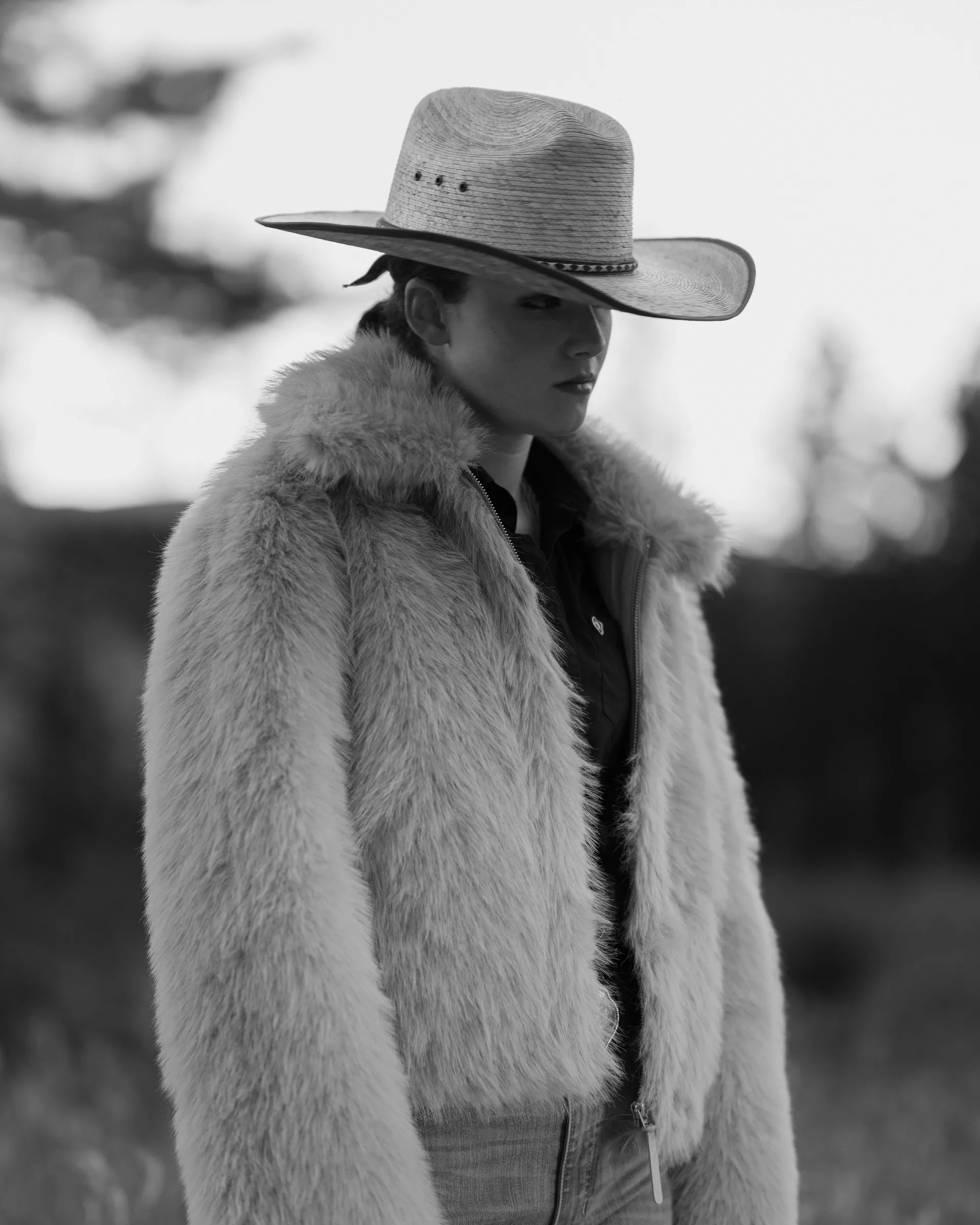 Black and white portrait of a woman in a straw cowboy hat and shaggy fur coat standing in an open field