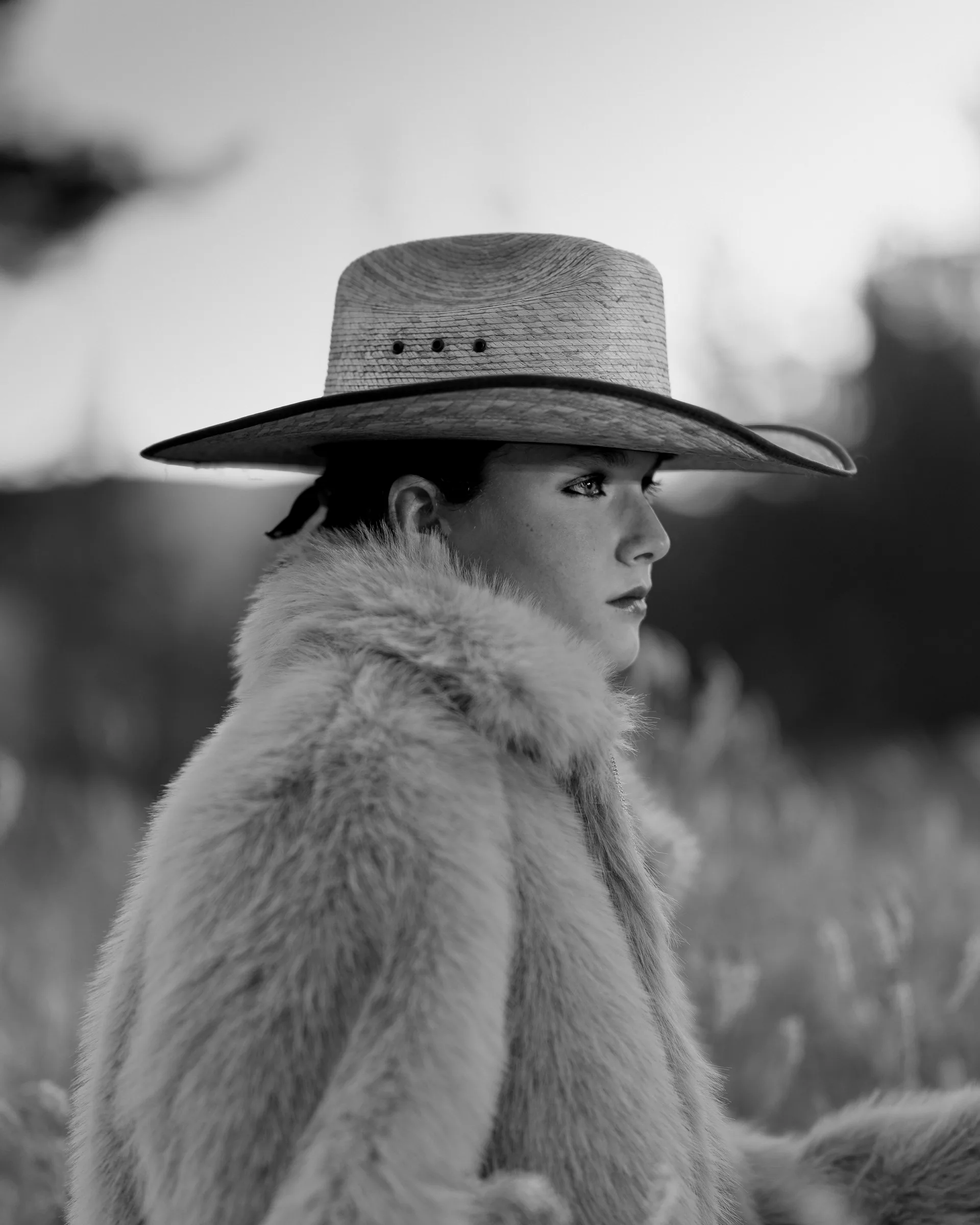 Black and white profile portrait of a woman in a straw cowboy hat and fur coat gazing into the distance in a field
