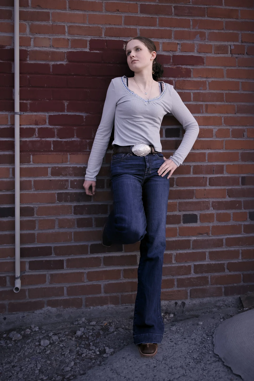 Woman in a grey top and flare jeans leaning against a red brick wall