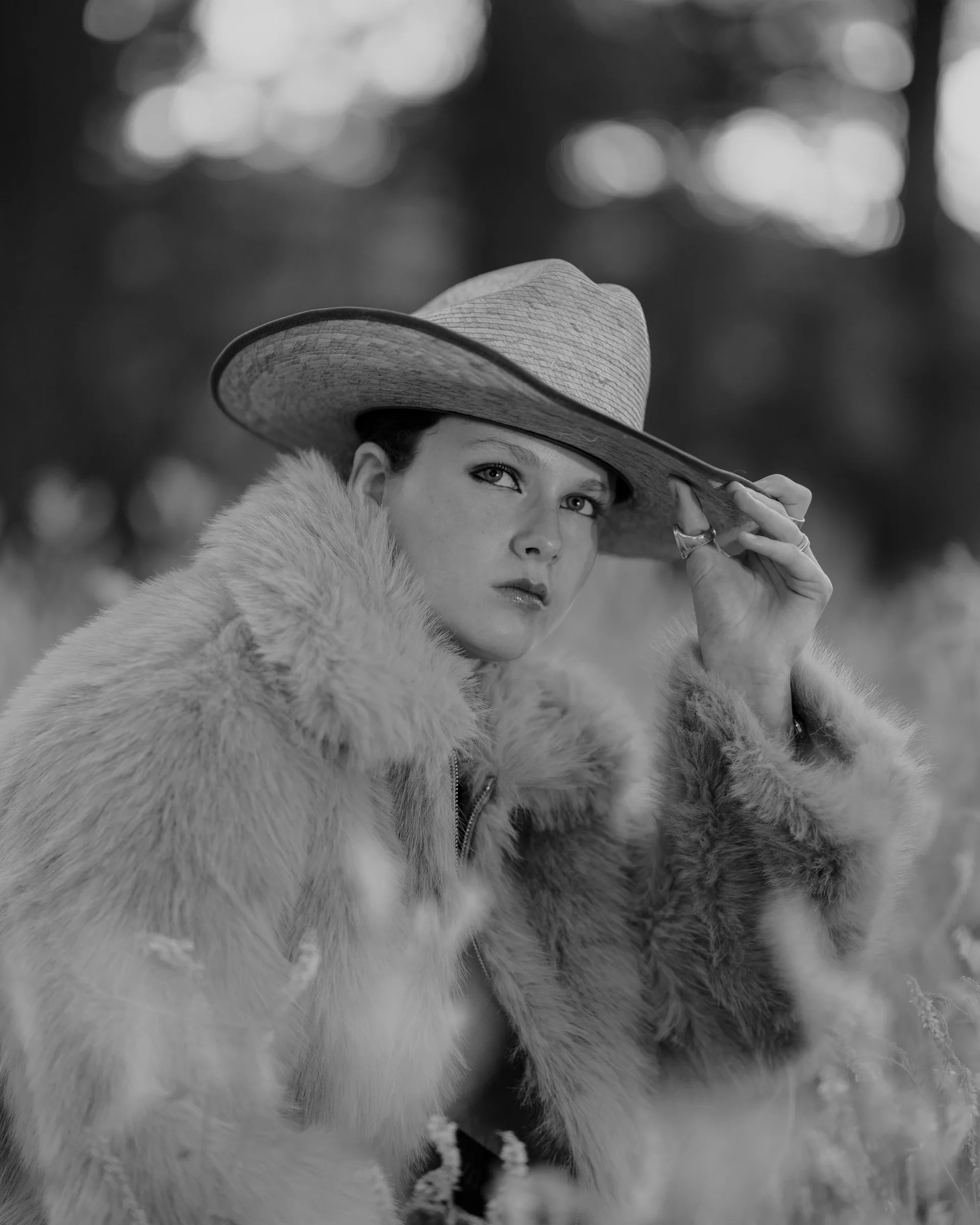 Black and white close-up portrait of a woman in a straw cowboy hat and fur coat reaching up to touch the brim