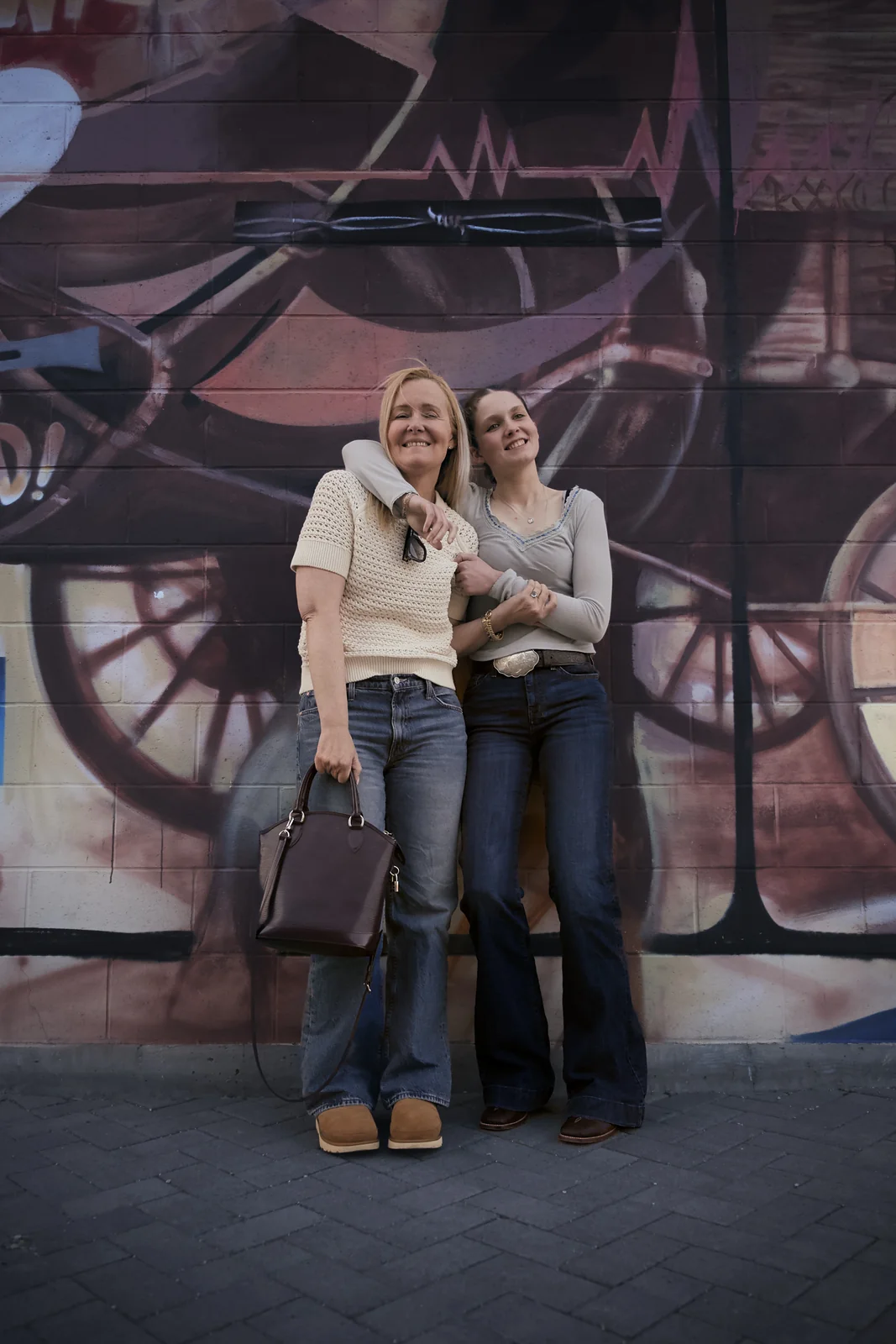 Two women embracing and smiling in front of a colorful graffiti mural on a city street