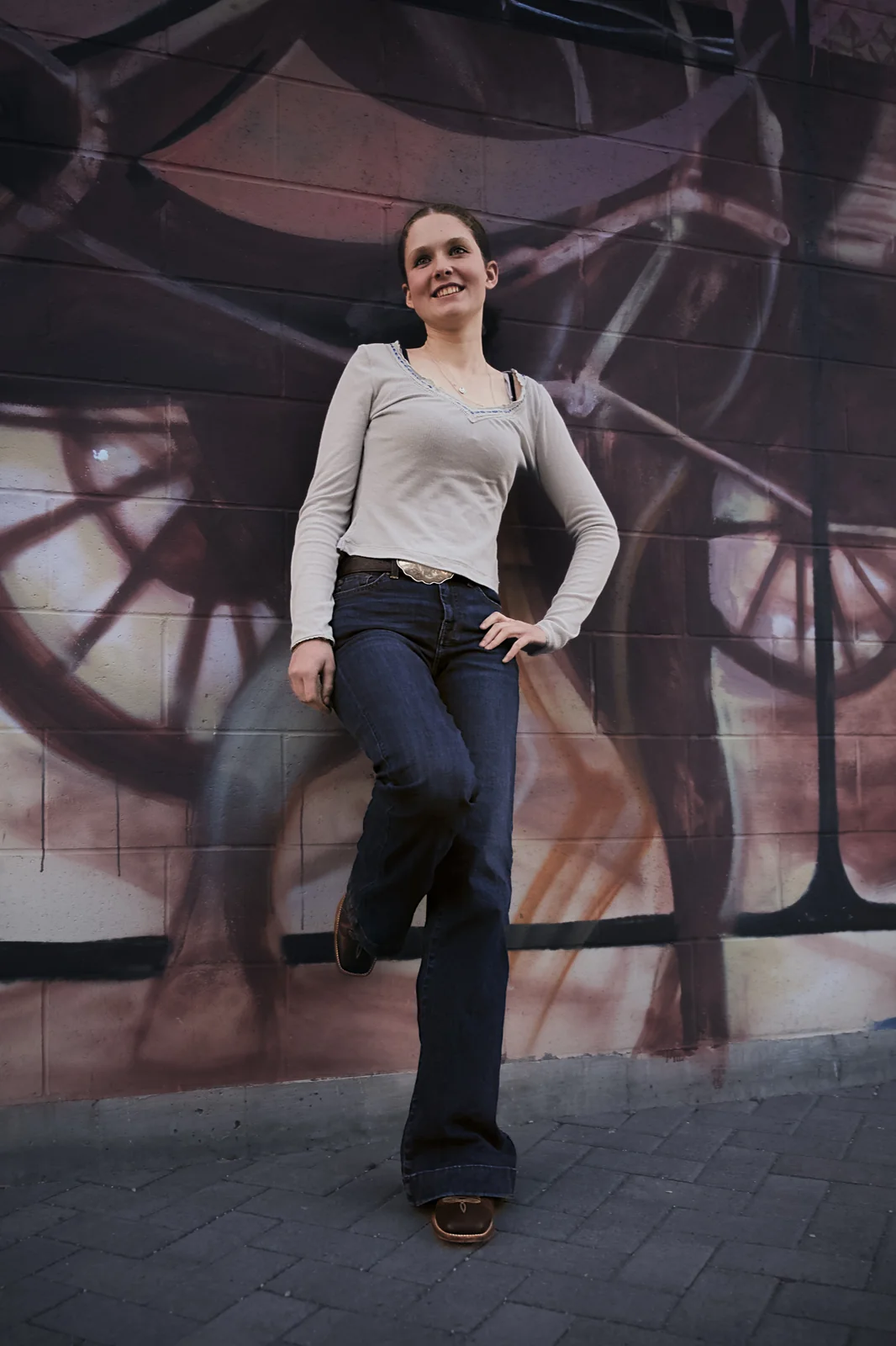 Woman in a grey top and flare jeans smiling and leaning against a graffiti wall