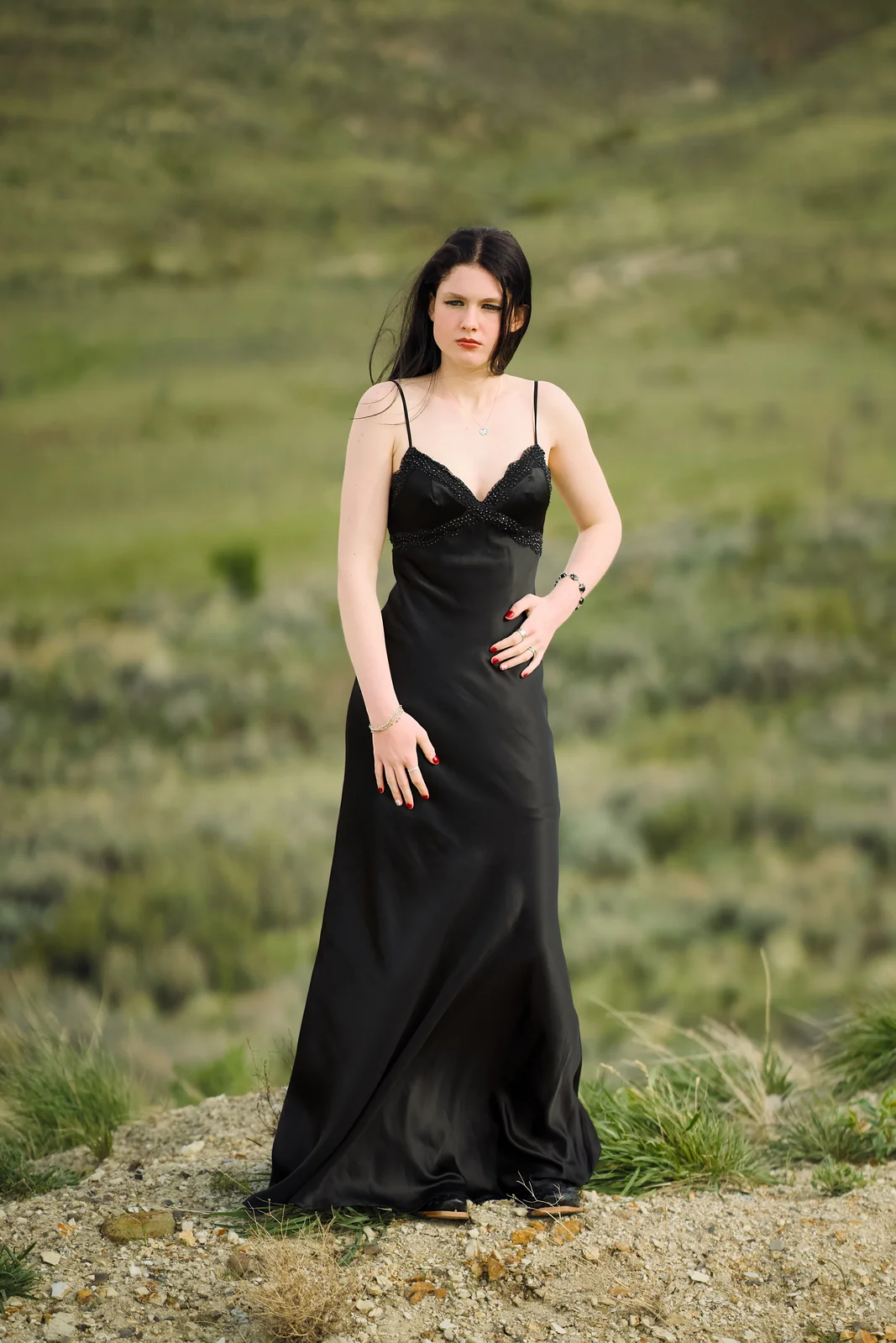 Woman in a flowing black satin gown standing in an open desert landscape with rolling green hills