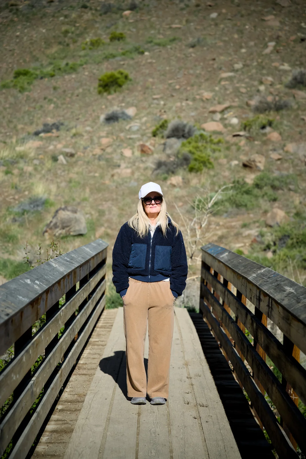 Woman in a navy fleece jacket and white cap standing on a wooden bridge with rocky hills behind