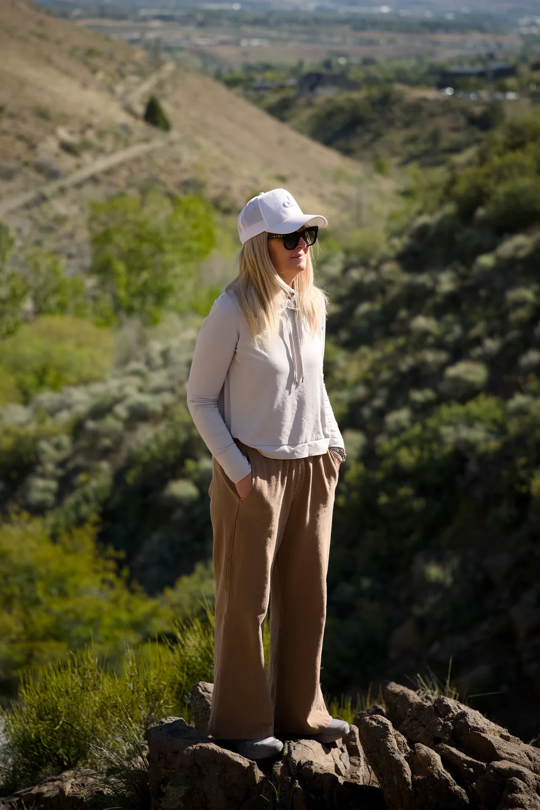 Woman in a grey hoodie and tan wide-leg pants standing on a rocky ridge with green hills in the background