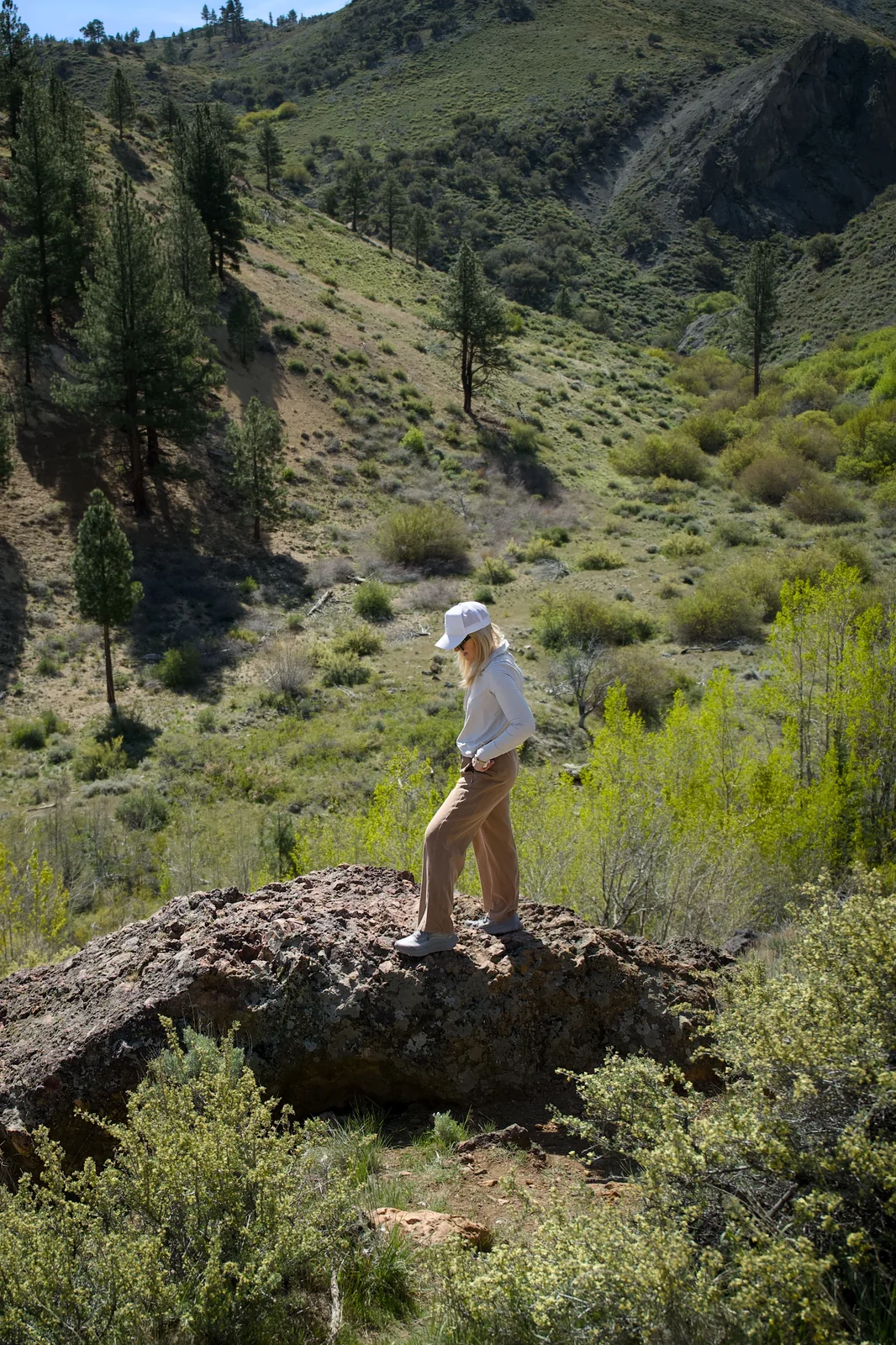 Woman in a white shirt and tan pants standing on a large boulder in a lush canyon landscape