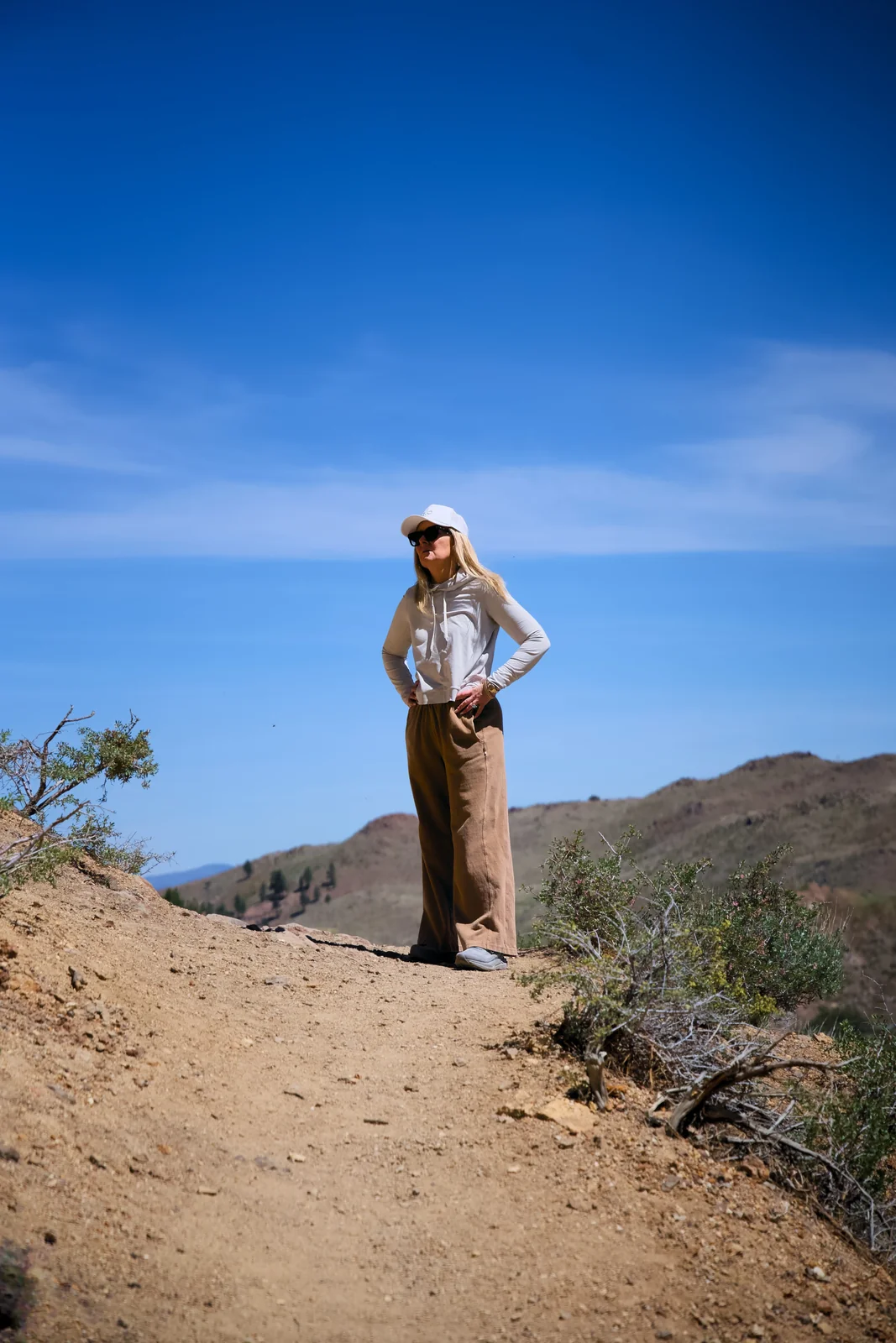 Woman in a light jacket standing on a dirt trail under a blue sky with arid hills behind