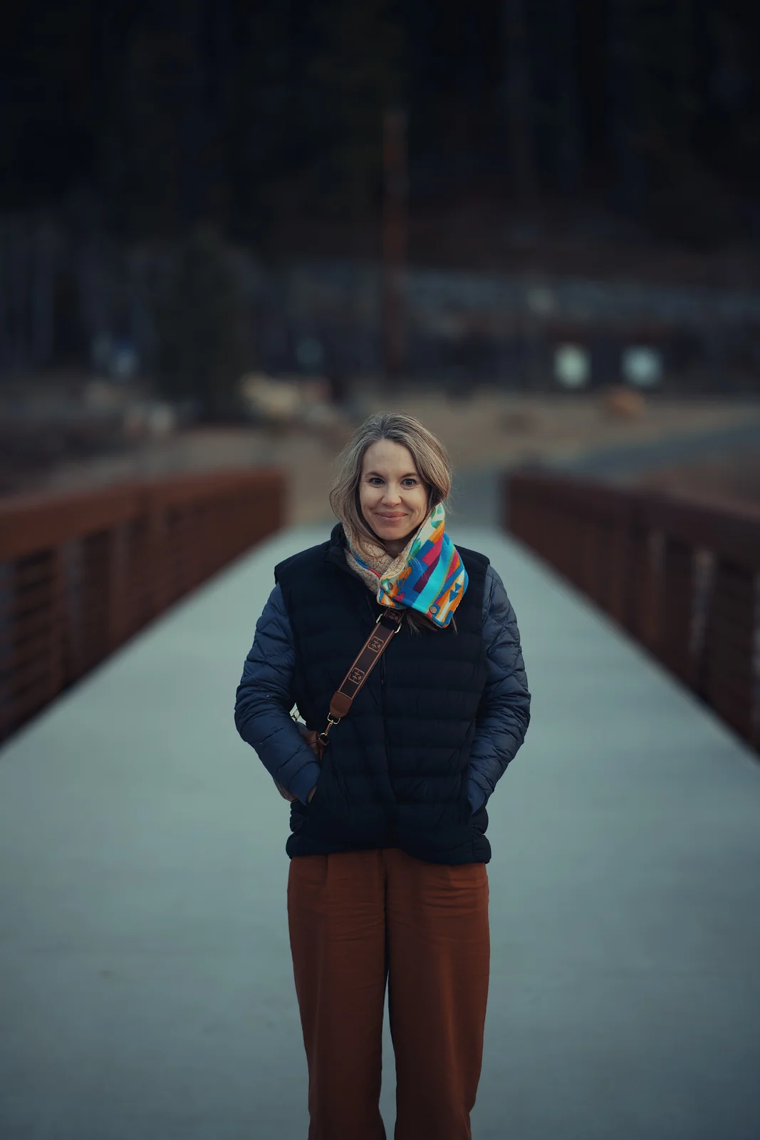 Woman smiling on a footbridge in a navy puffer vest and colorful Pendleton scarf