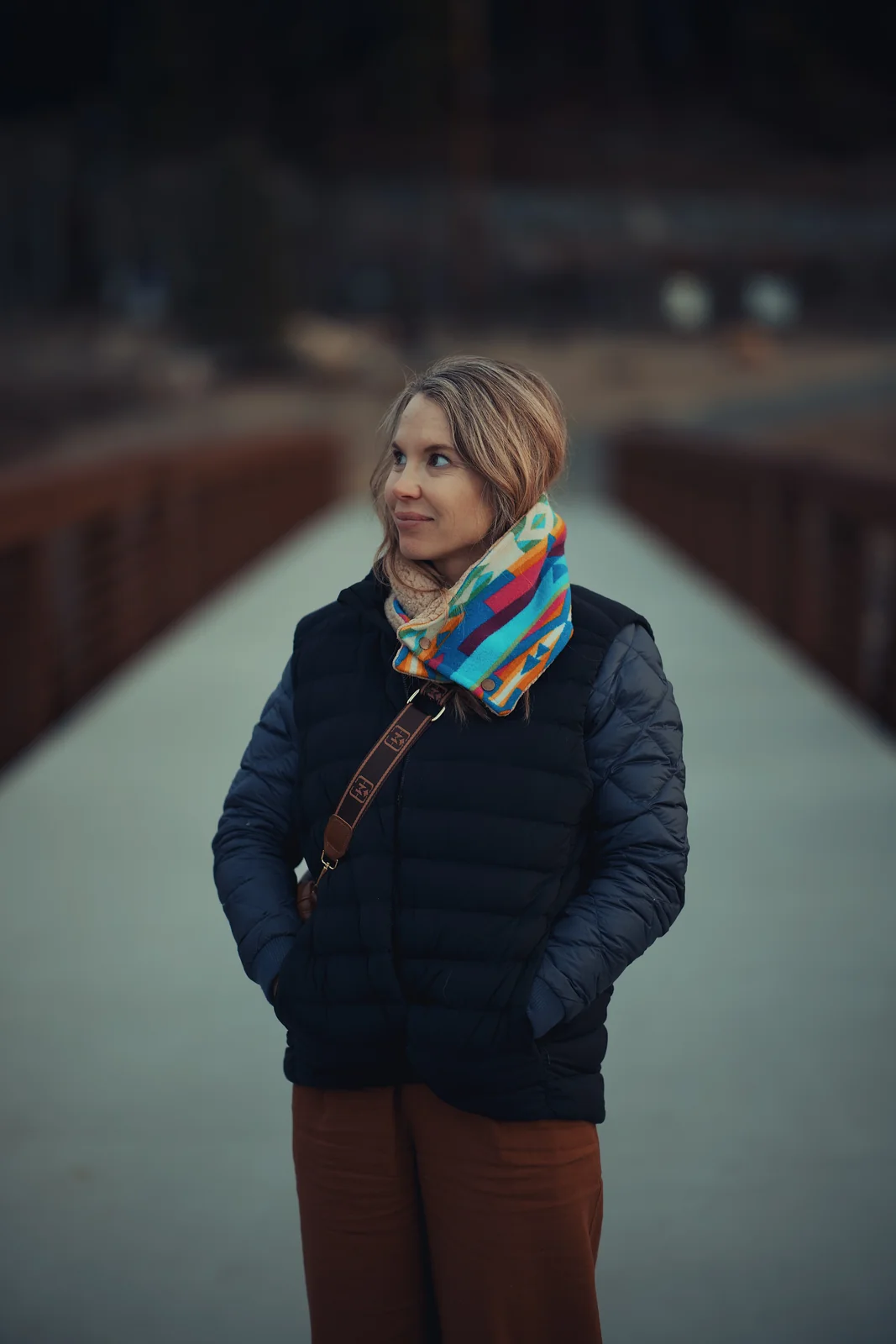 Woman in a navy puffer vest and colorful Pendleton scarf glancing to the side on a footbridge
