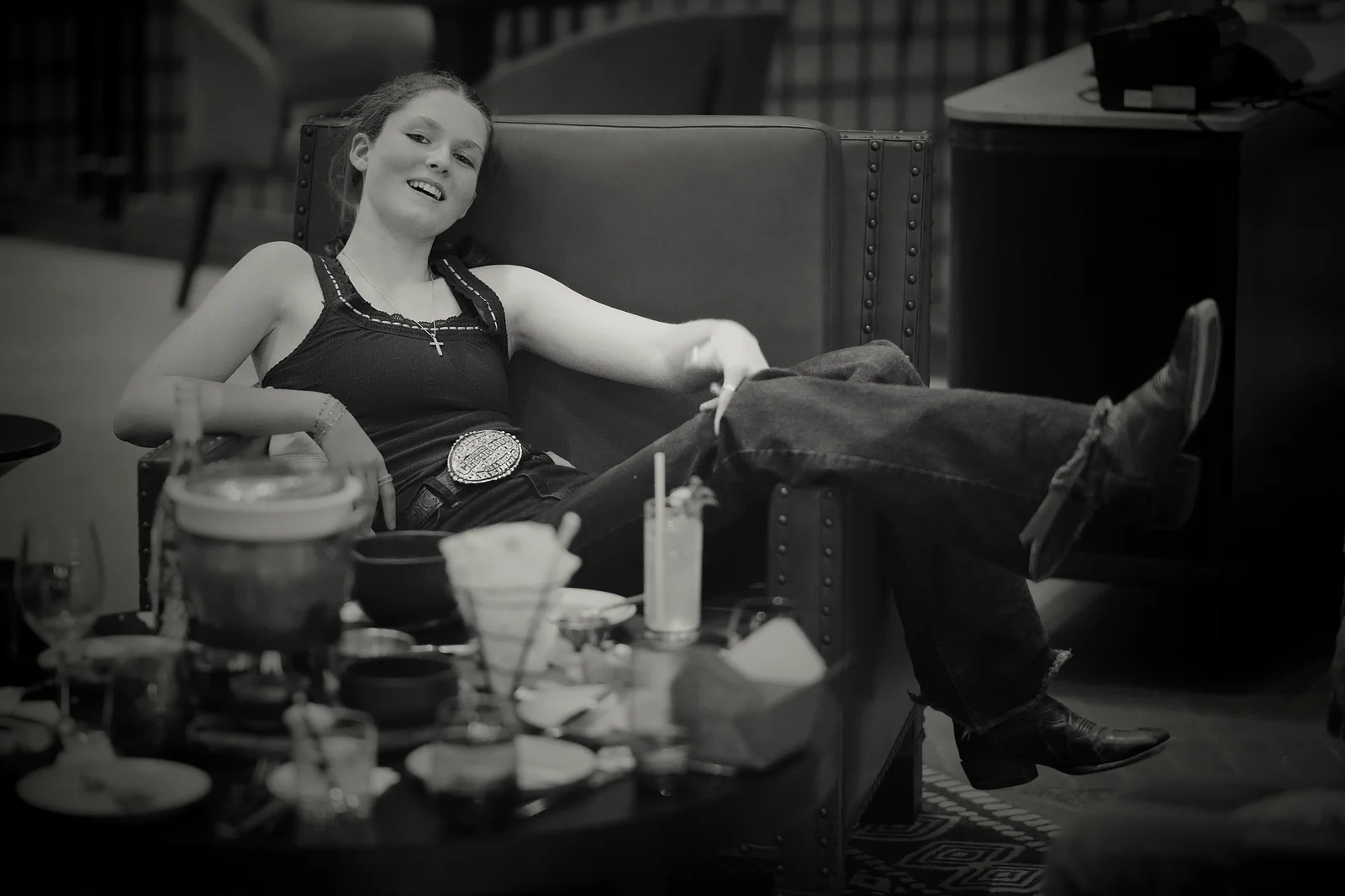 Young woman in a black tank top with a cowboy belt buckle sitting relaxed in an armchair, black and white