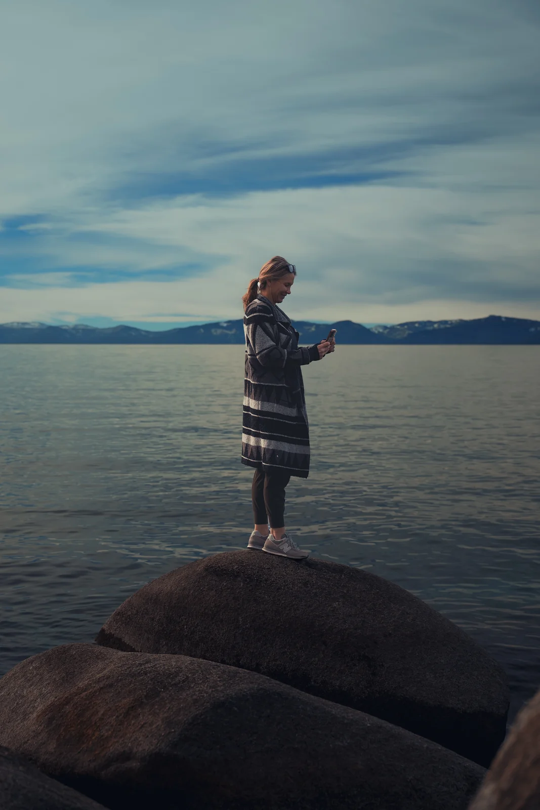 Woman in a striped coat standing on a large boulder at the edge of a calm lake with mountains behind